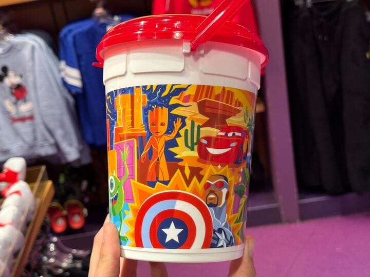 A vibrant popcorn bucket adorned with playful cartoon characters, including a tree, a car, and a superhero with a shield, sits cheerfully on a shelf next to merchandise in the store.