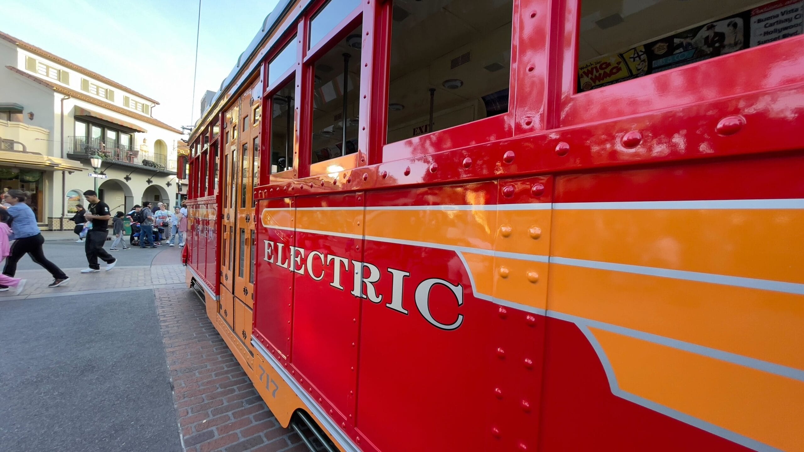Close-up of a bright red electric trolley with yellow accents on a city street. Dubbed the Red Car Trolley, it brings to mind the charm of Disney California Adventure. People stroll nearby, and buildings serve as a charming backdrop.