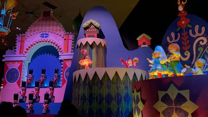 Colorful indoor diorama featuring whimsical figures, including dolls and toy soldiers, evokes the charm of Magic Kingdom's "it's a small world," set against a backdrop of bright, decorative structures.