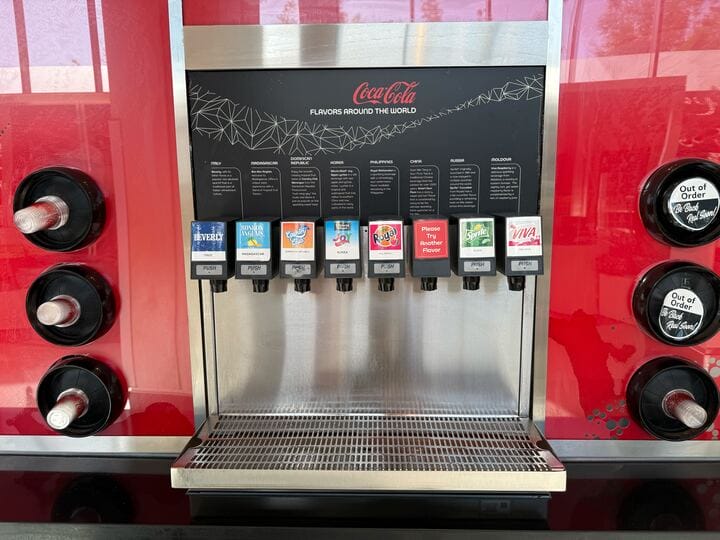 A soda fountain at Club Cool, featuring eight beverage options including various Coca-Cola flavors and unique selections like Sour Plum Tea. Two out-of-order buttons are visible on the sides.