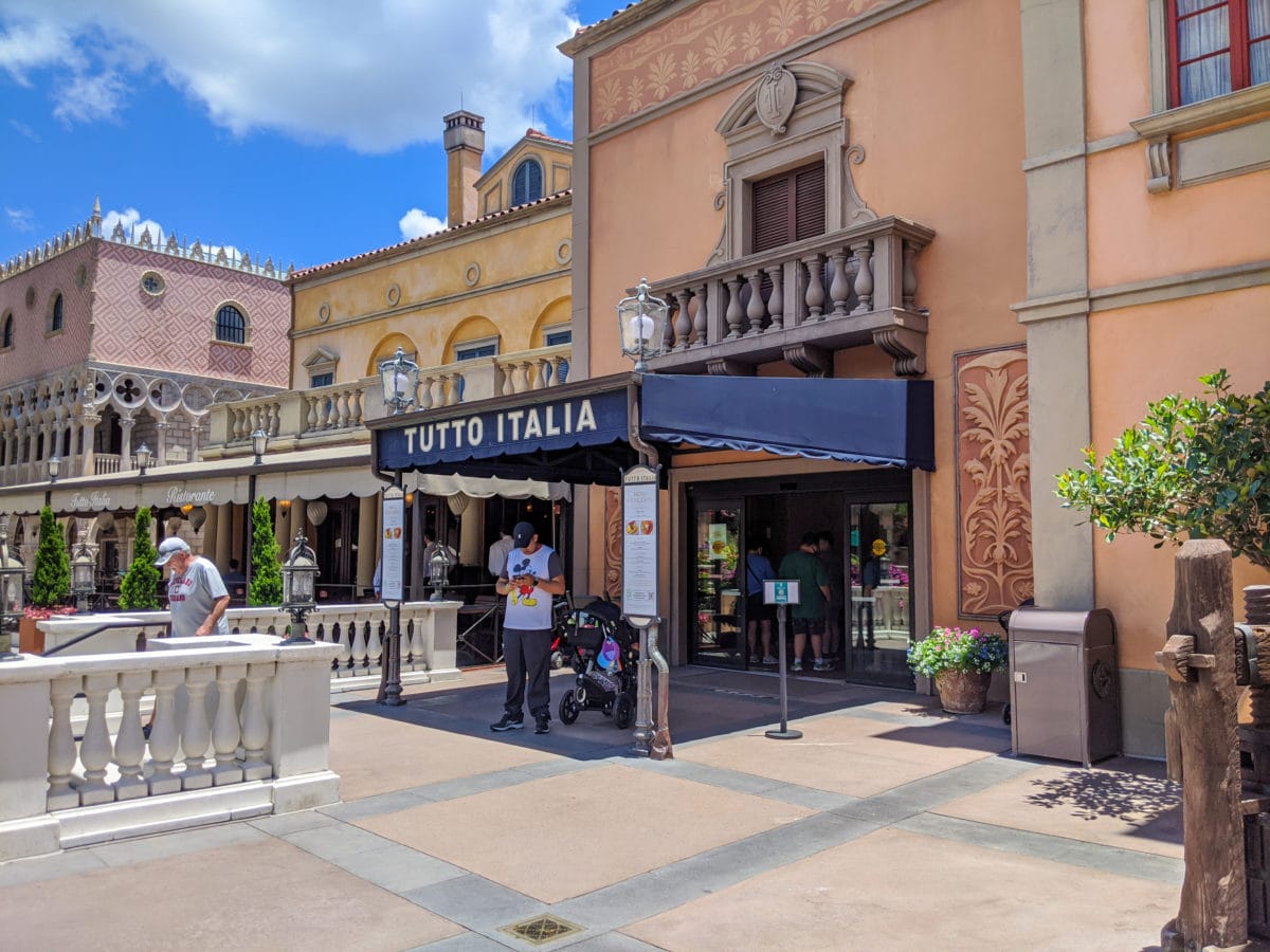 The exterior of Tutto Italia restaurant at EPCOT showcases a stylish black awning under a clear blue sky, with visitors leisurely standing outside.