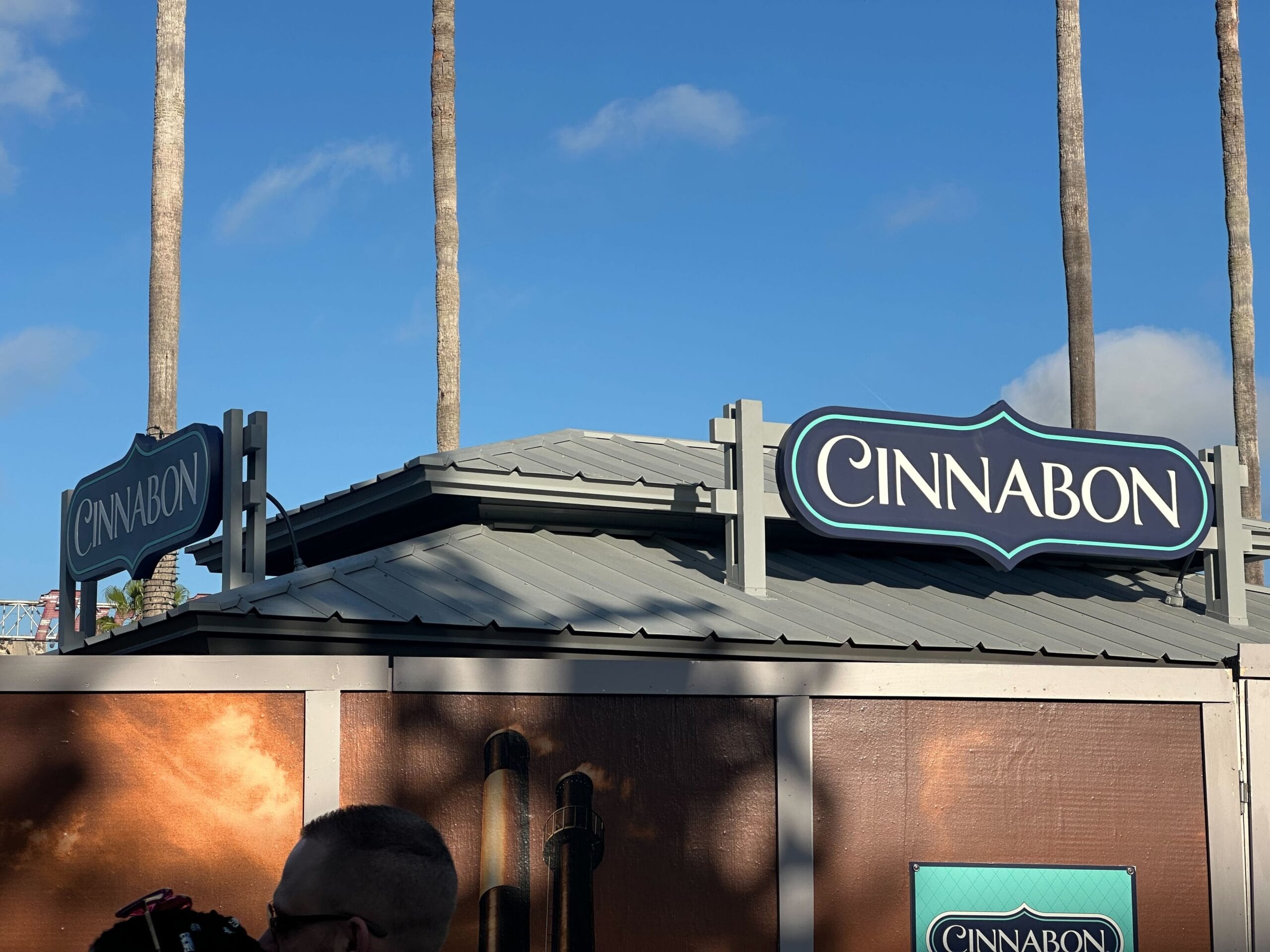 Cinnabon store with signs on a gray roof, palm trees and blue sky in the background.