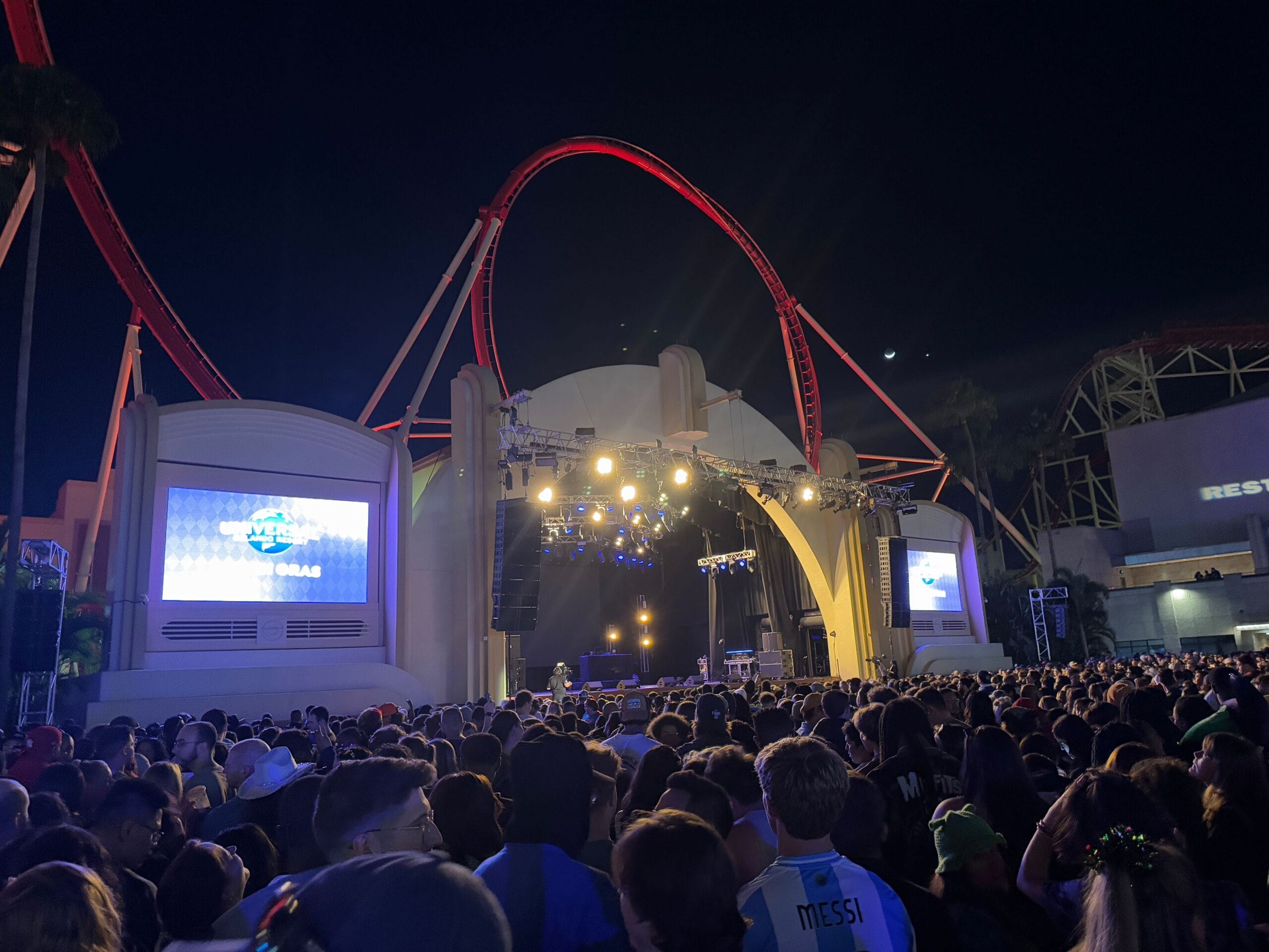 A large crowd gathered at an outdoor concert venue at night, featuring bright stage lights and a roller coaster in the background, as T-Pain brought his energy to the Universal Mardi Gras stage.