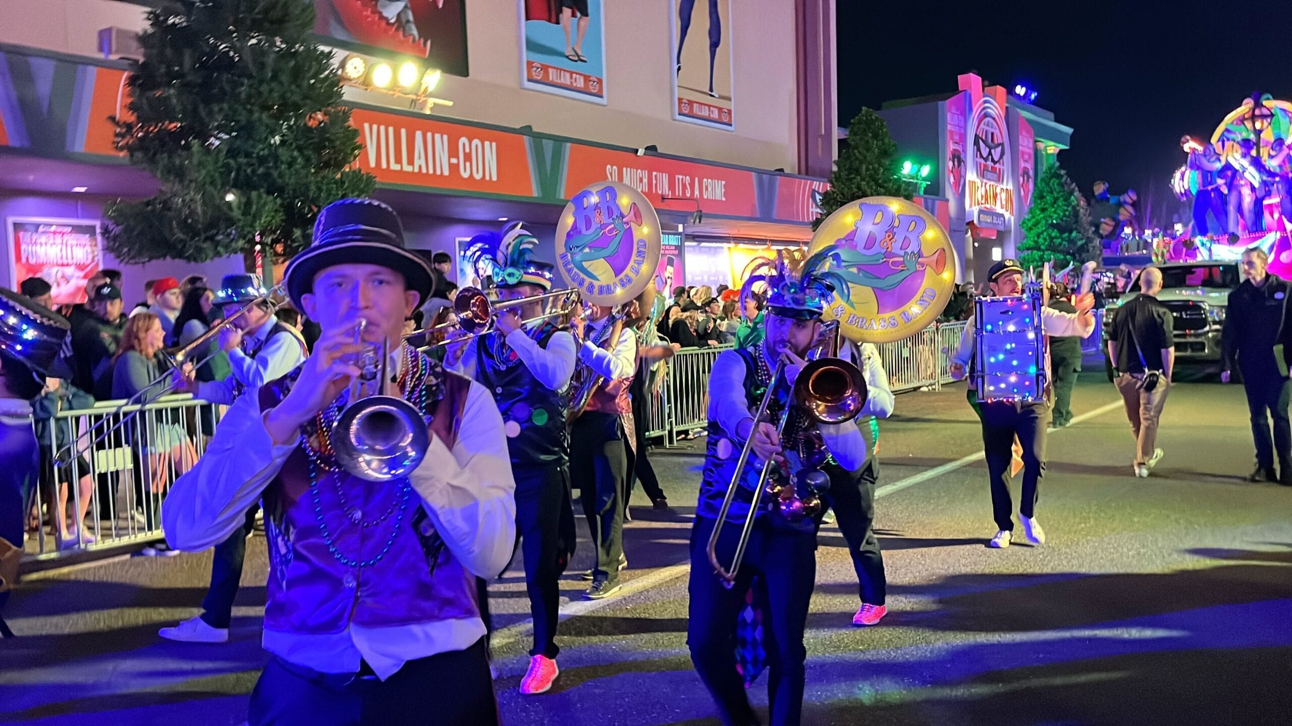 In the Universal Mardi Gras parade, a marching band in festive attire performs under the night sky, with musicians playing trombones and sousaphones. Bright lights and a colorful float illuminate the background, adding to the vibrant spectacle.
