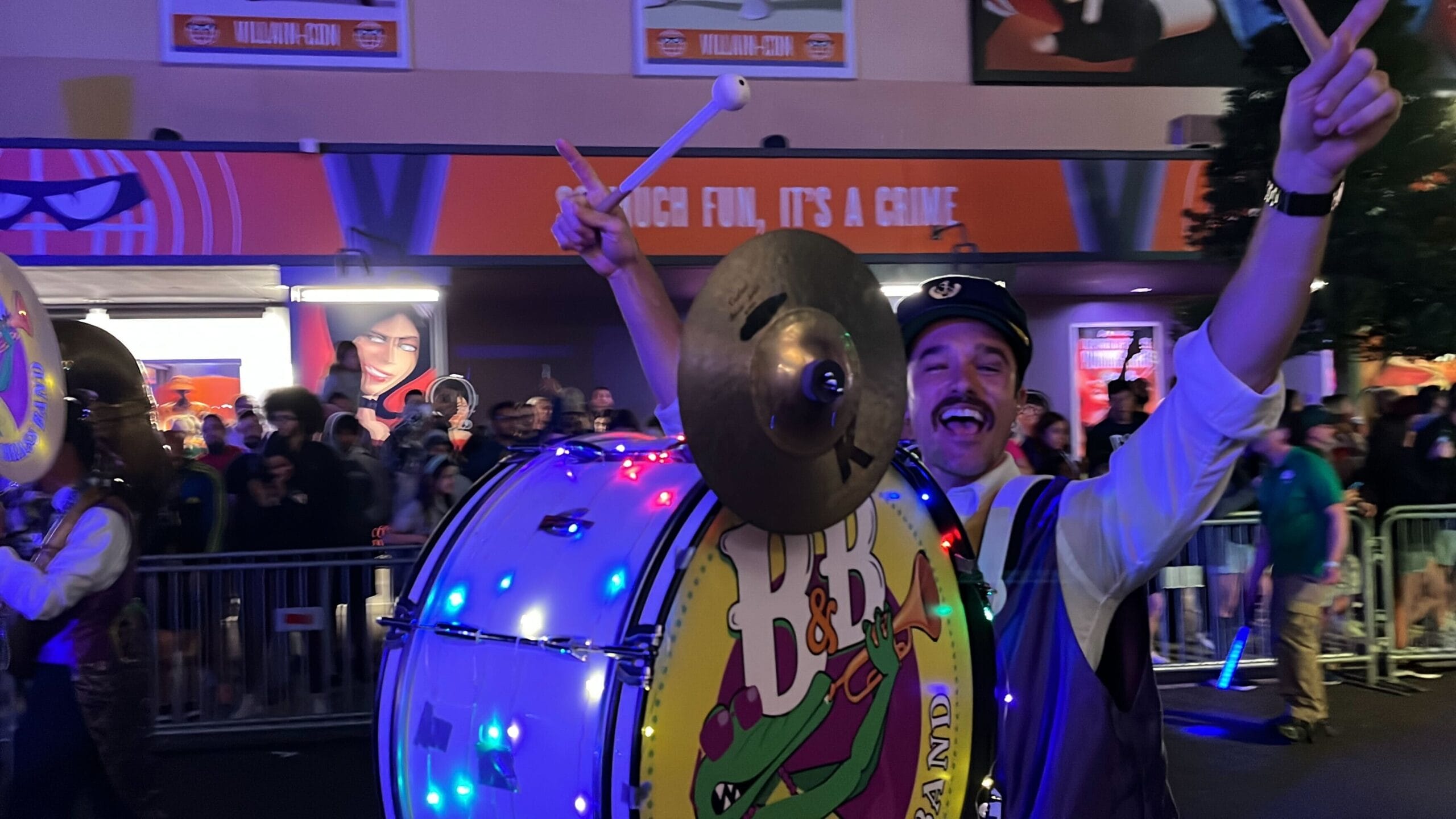 A man in the Universal Mardi Gras parade smiles widely while playing a bass drum adorned with lights. He holds a cymbal and drumstick in each hand, captivating the crowd watching behind him.