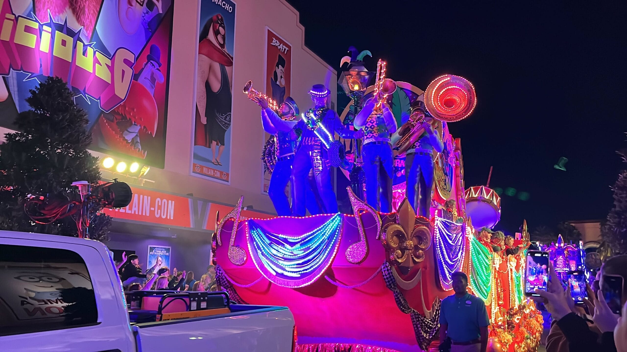 A colorful Universal Mardi Gras parade float, brimming with musicians and vibrant lights, glides through a crowd-lined street at night.