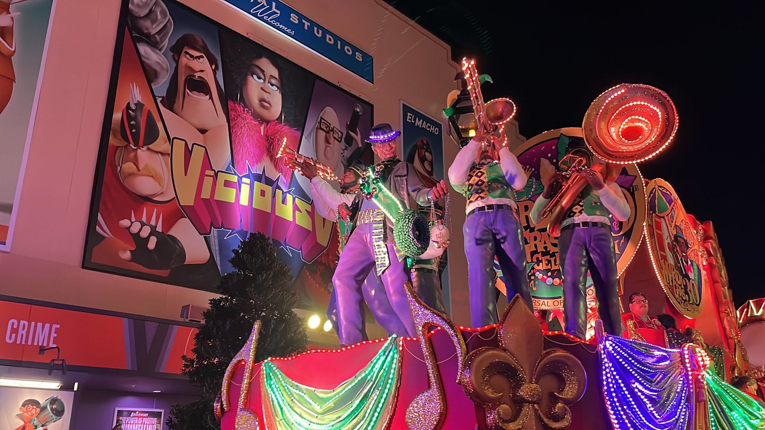 A colorful parade float with musicians and festive lights glides by a large building adorned with animated movie posters, capturing the lively spirit of the Universal Mardi Gras parade.