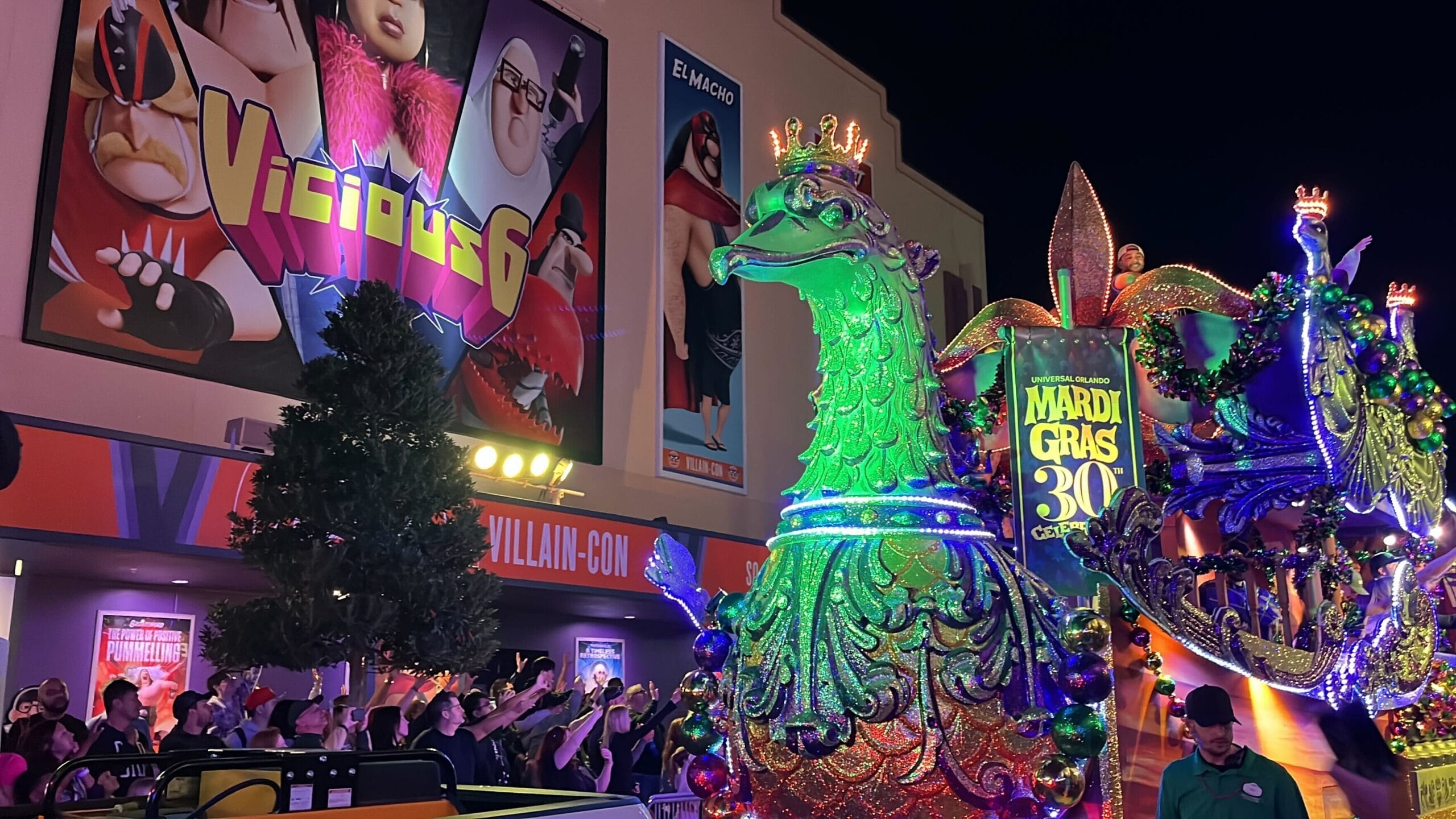 A colorful Mardi Gras float from the Universal Mardi Gras parade showcases a lit-up peacock with a crown, gliding past a lively crowd. In the background, a movie theater displays its latest attraction: the "Vicious 6" poster.