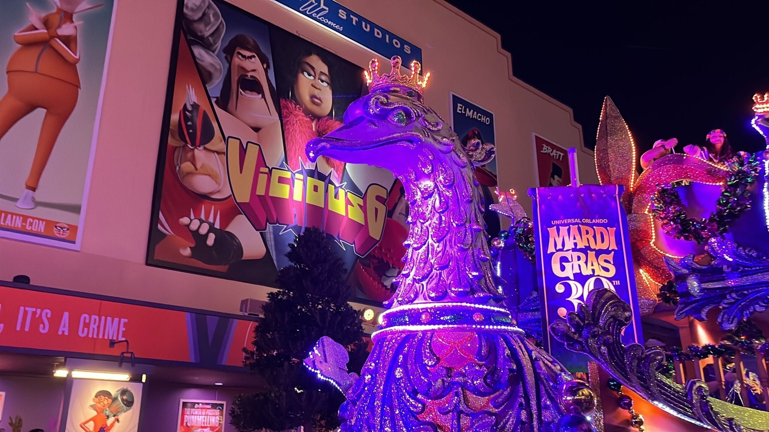 A colorful Mardi Gras float featuring a large, ornate purple bird makes its grand appearance in the Universal Mardi Gras parade, set against a vibrant backdrop of theme park posters.