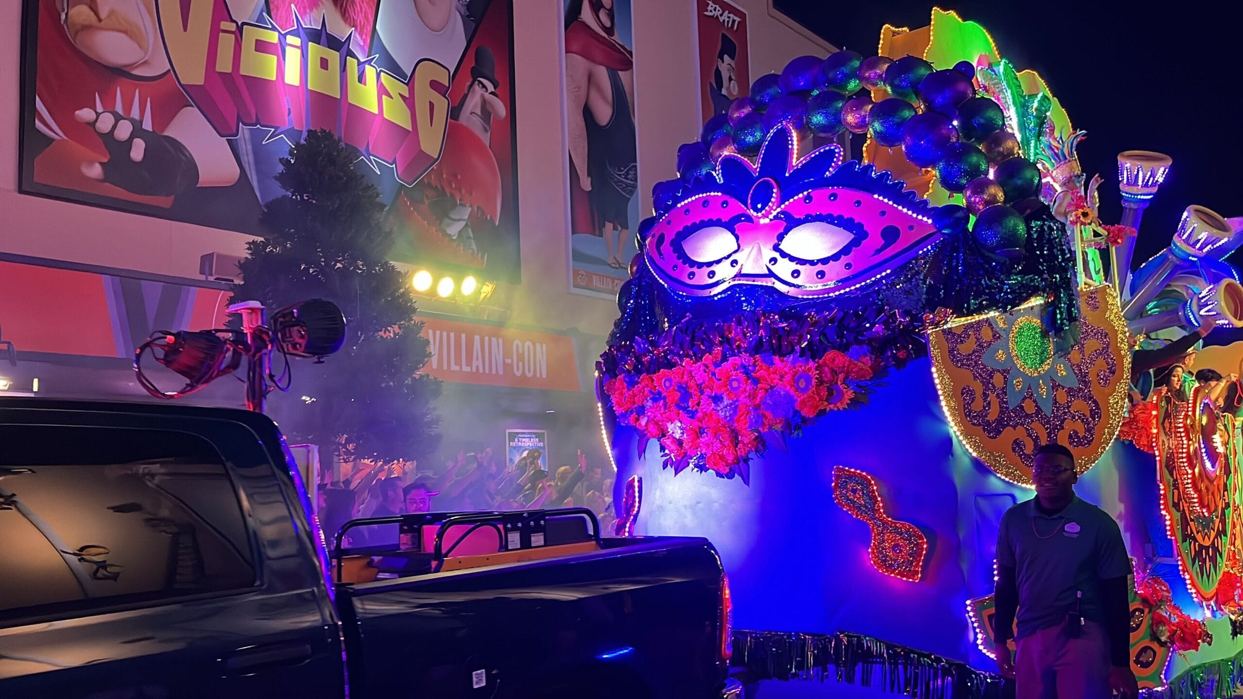 A parade float, part of the Universal Mardi Gras parade, is adorned with a large, colorful mask and vibrant decorations, glowing beautifully in the night. A person stands nearby.