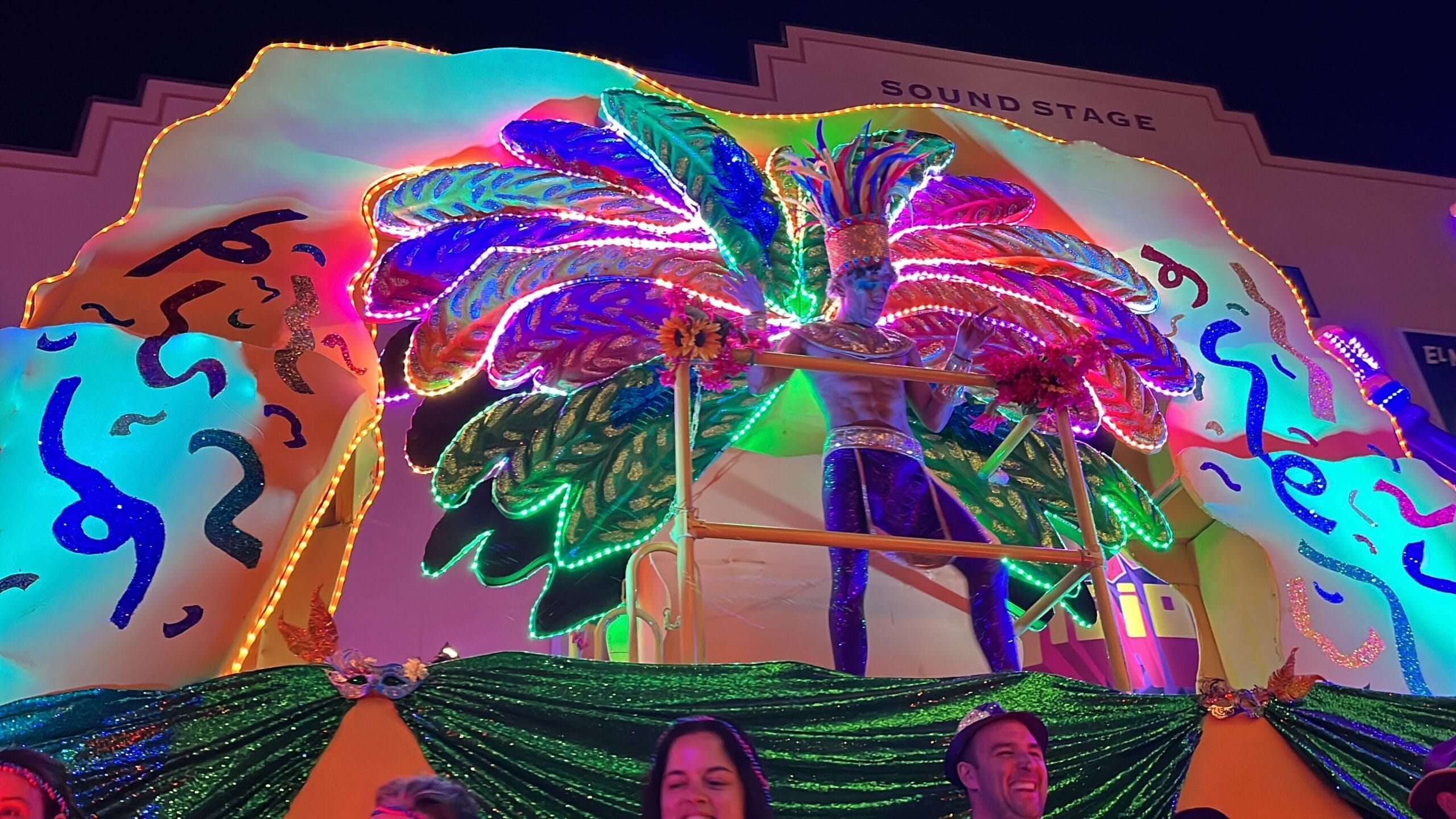 The Universal Mardi Gras parade features a vibrant float with a performer in a feathered costume and dazzling neon lights. Enthusiastic onlookers are smiling, captivated by the lively spectacle.
