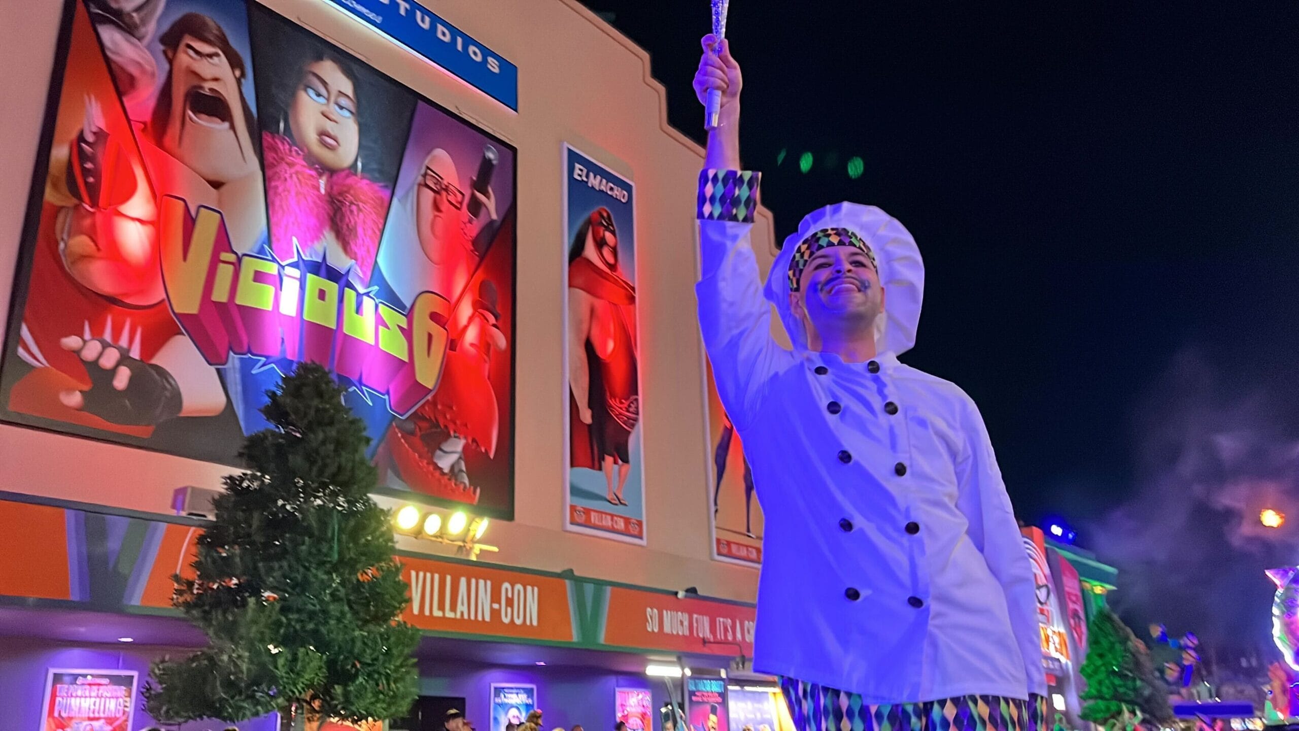 A performer in a chef costume, whisk held high, dazzles in front of a building adorned with animated villain posters during the vibrant Universal Mardi Gras parade at night.