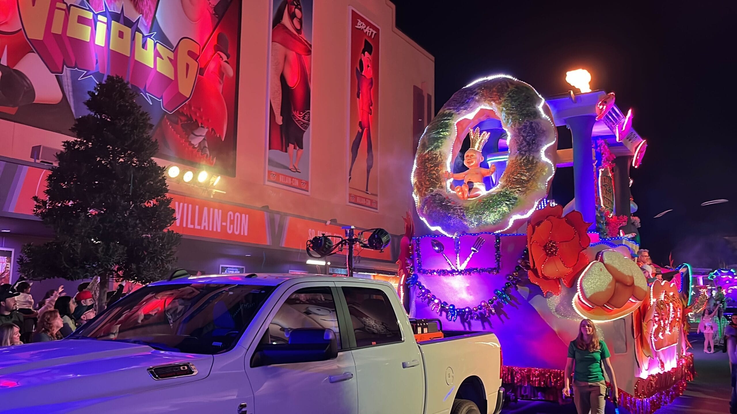 A brightly illuminated parade float adorned with large flowers and a wreath is towed by a white truck at night, embodying the universal spirit of Mardi Gras. A person waves jubilantly from the float as spectators eagerly watch the vibrant procession pass by.