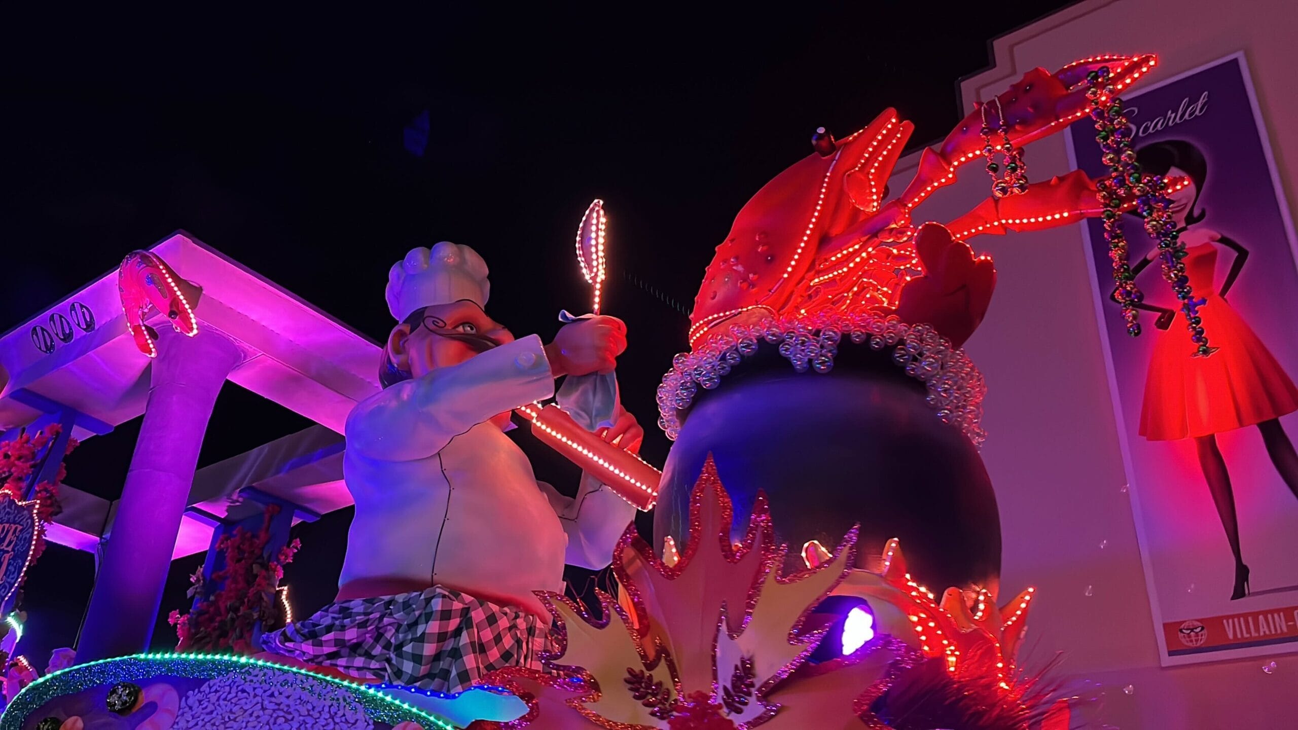 A colorful parade float at the Universal Mardi Gras parade features a chef with fork and knife poised, facing off against a large crab in a pot. Bright neon lights and vivid decorations bring this culinary showdown to life.