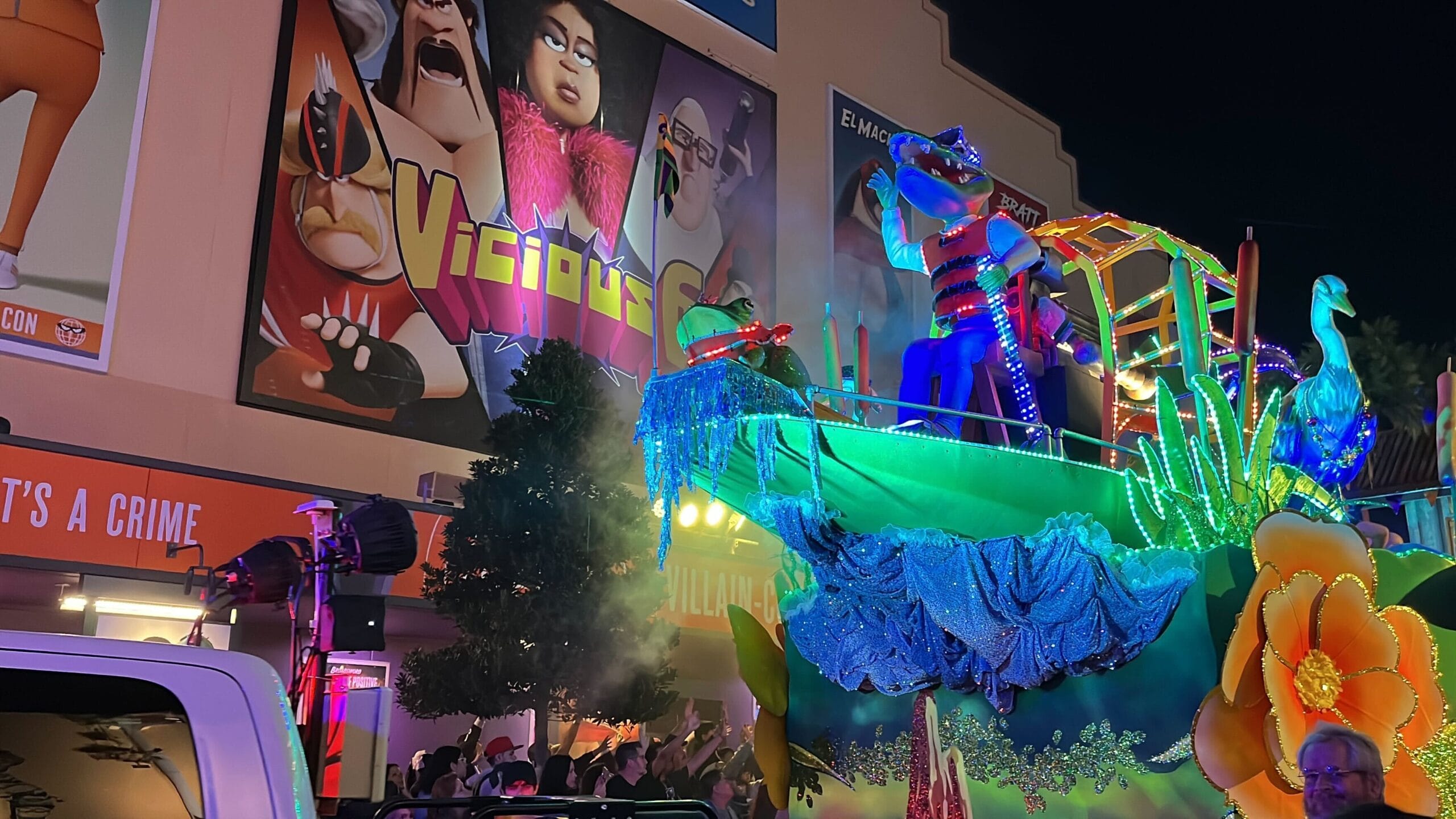 A vibrant Universal Mardi Gras parade float, adorned with dazzling lights and decorations, glides through the street under the night sky, enthralling a lively crowd. Large animated character posters enhance the festive atmosphere in the background.