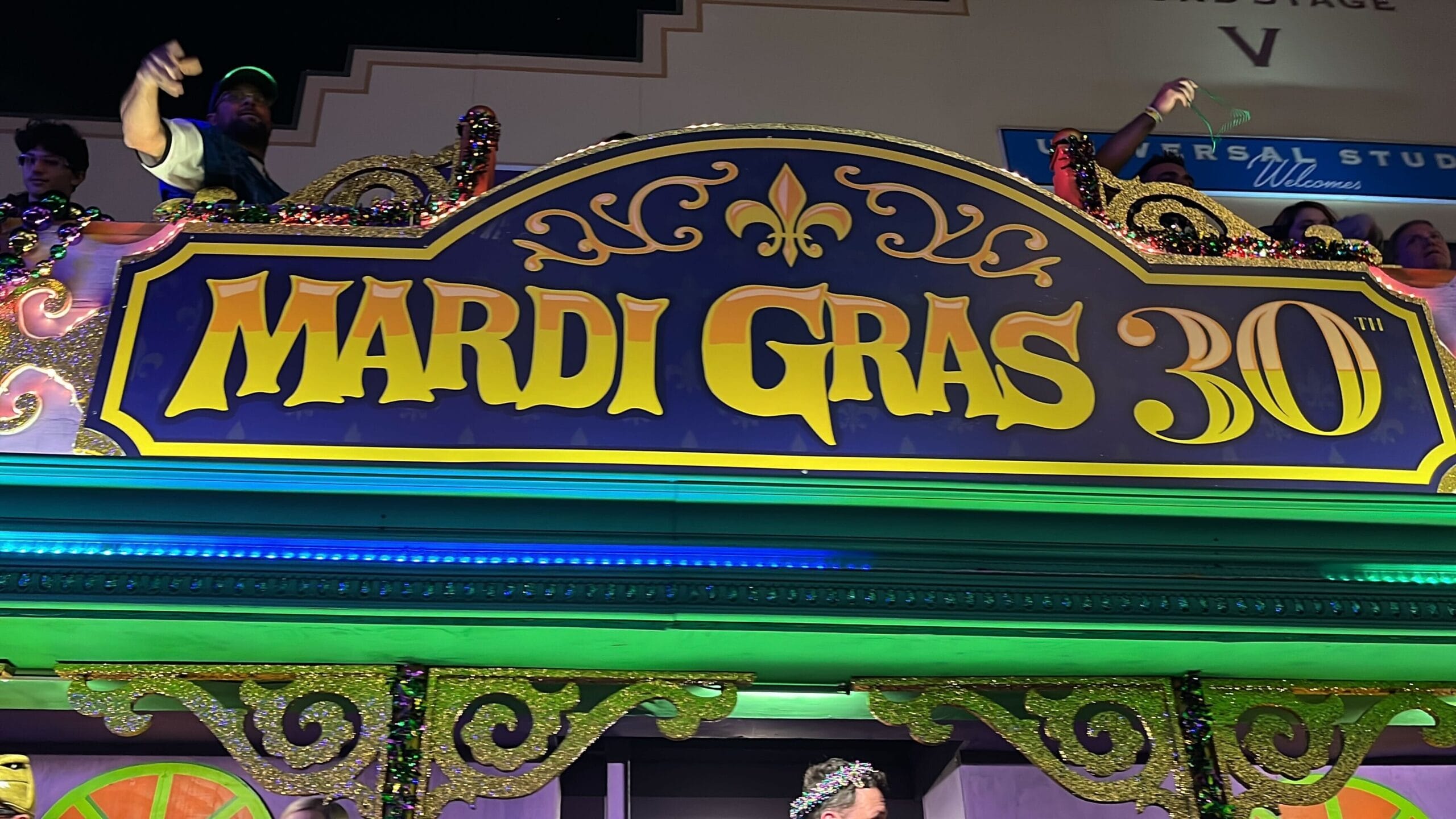 A colorful sign reads "Mardi Gras 30th" with ornate decorations, including a fleur-de-lis, capturing the spirit of the Universal Mardi Gras parade against a backdrop of people on an upper level.