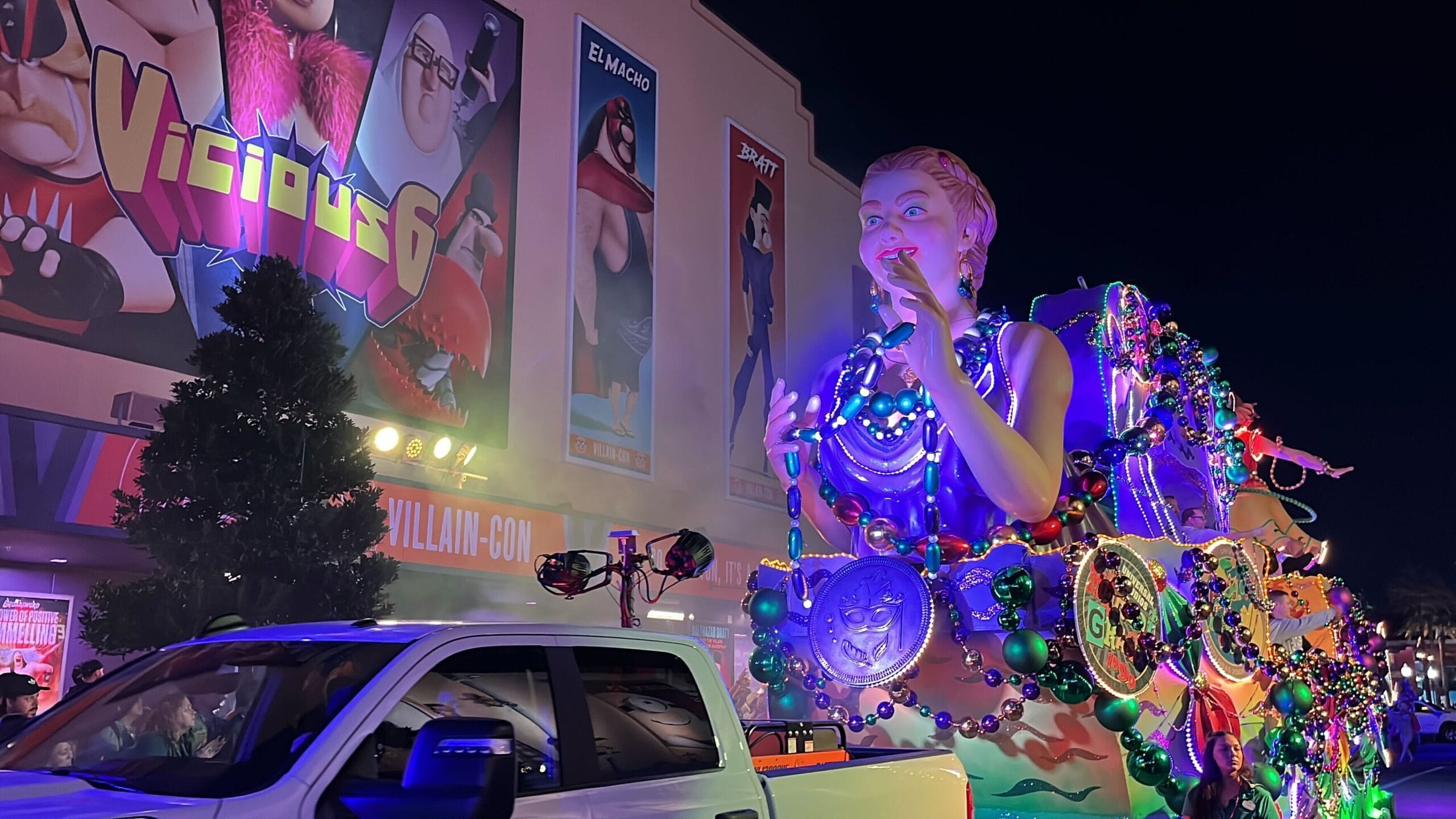 A Universal Mardi Gras parade float featuring a large, colorful figure adorned with beads and medallions glides beside a white truck in front of an illuminated building with cartoon movie posters at night.