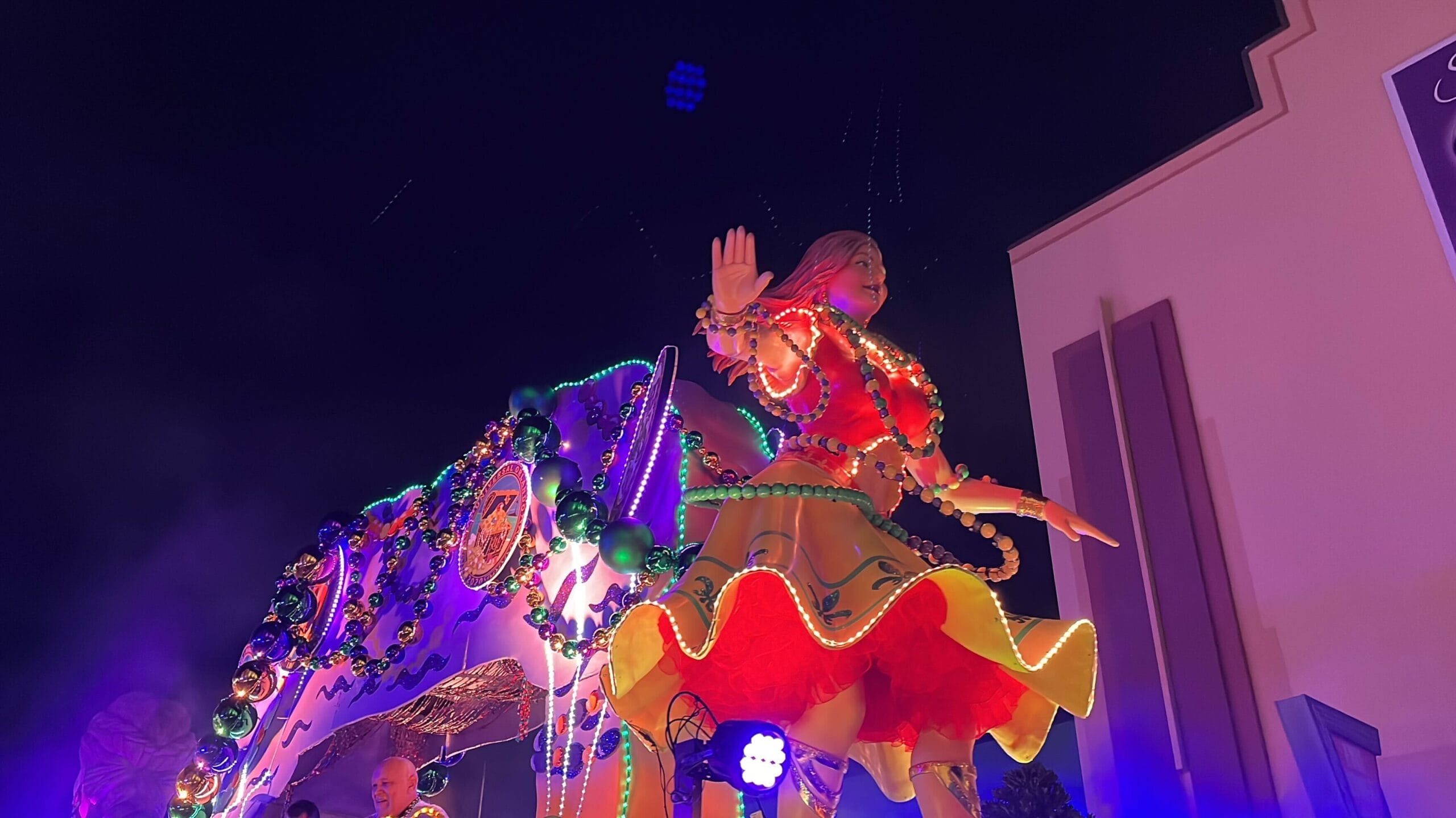 A brightly lit parade float at the Universal Mardi Gras parade features a large, colorful statue of a person in festive attire with lights, set against the dark night sky.