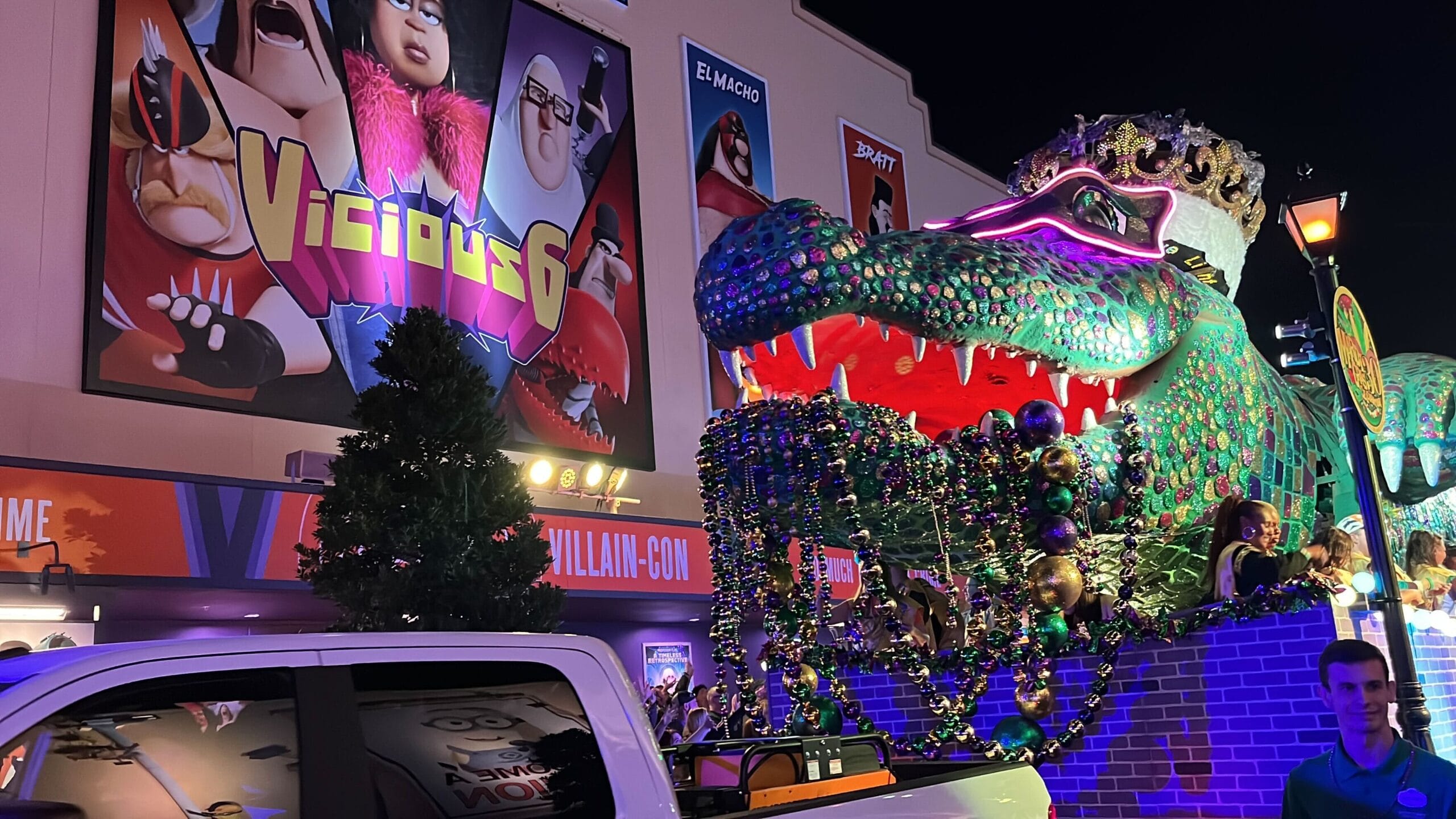 The Universal Mardi Gras parade dazzles under the night sky with a large float showcasing a colorful alligator adorned with beads. Billboards in the background come alive, featuring animated characters and text that reads "Vicious 6," adding an extra layer of excitement to the festivities.