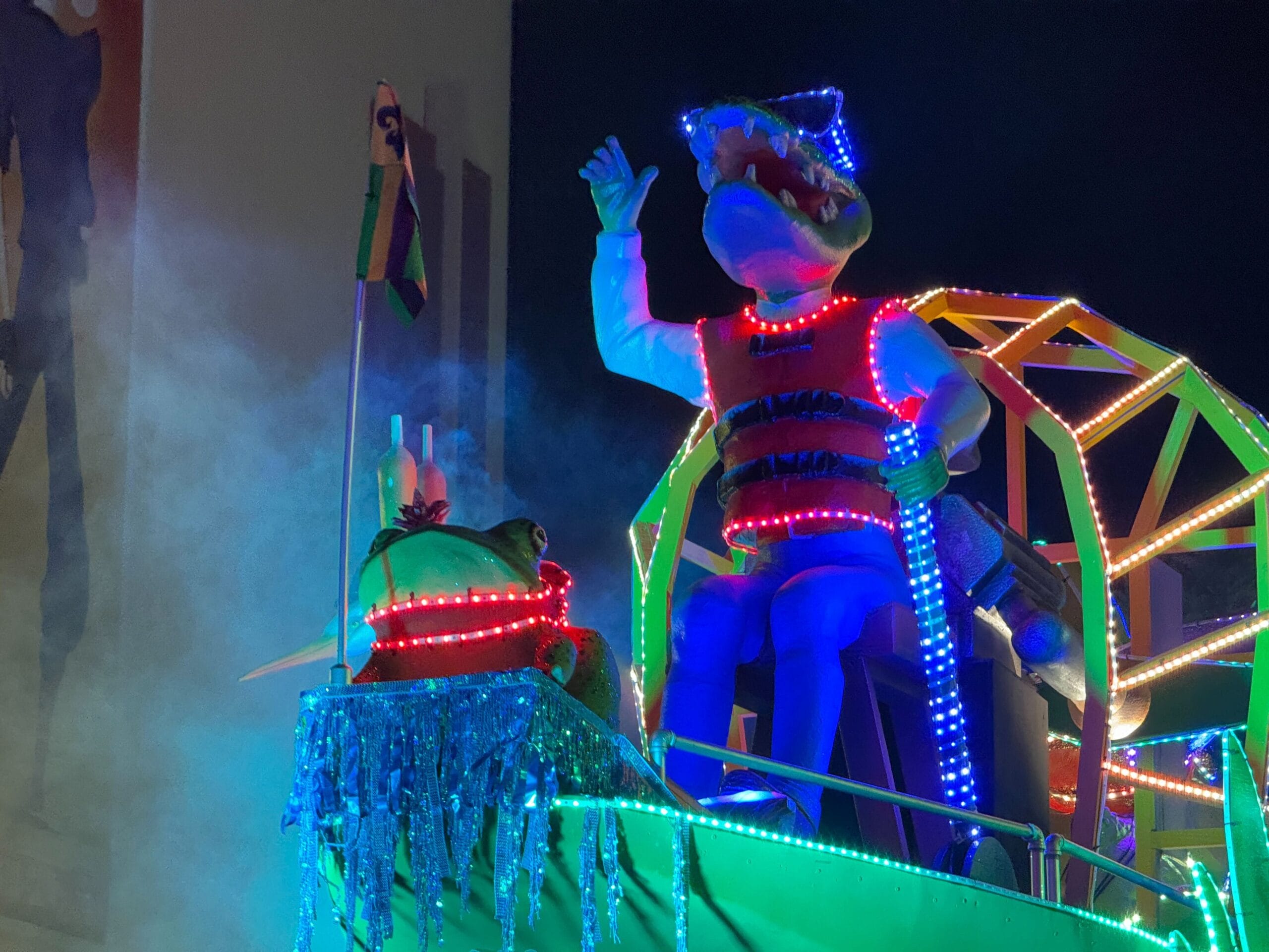 An illuminated alligator costume figure, a highlight of the Universal Mardi Gras parade, sits on a colorful float surrounded by lights and fog, waving a glowing wand.