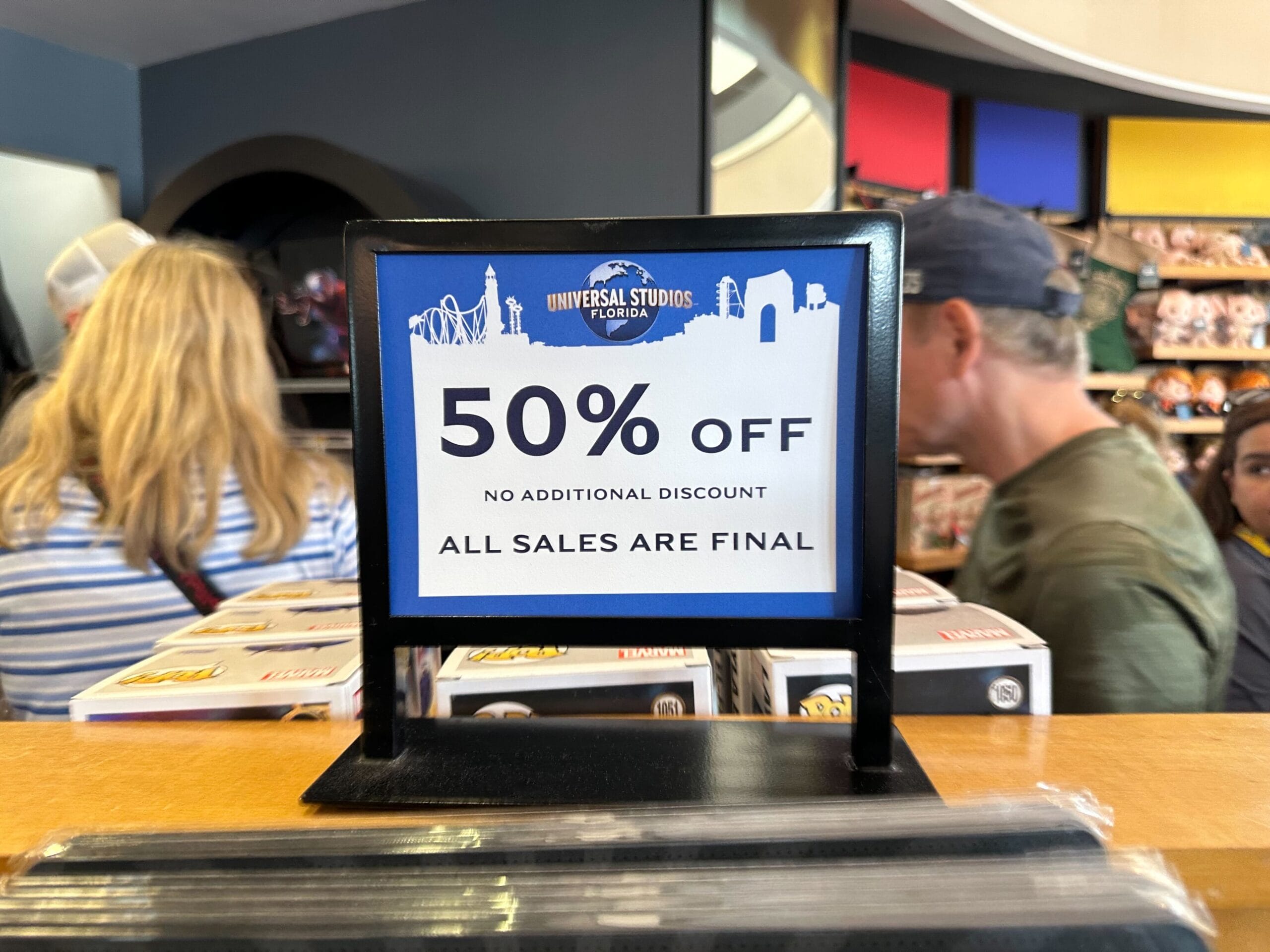 Sign at a shop counter reading "50% Off No Additional Discount All Sales Are Final" with a Universal Studios Florida logo. Customers and merchandise are visible in the background.