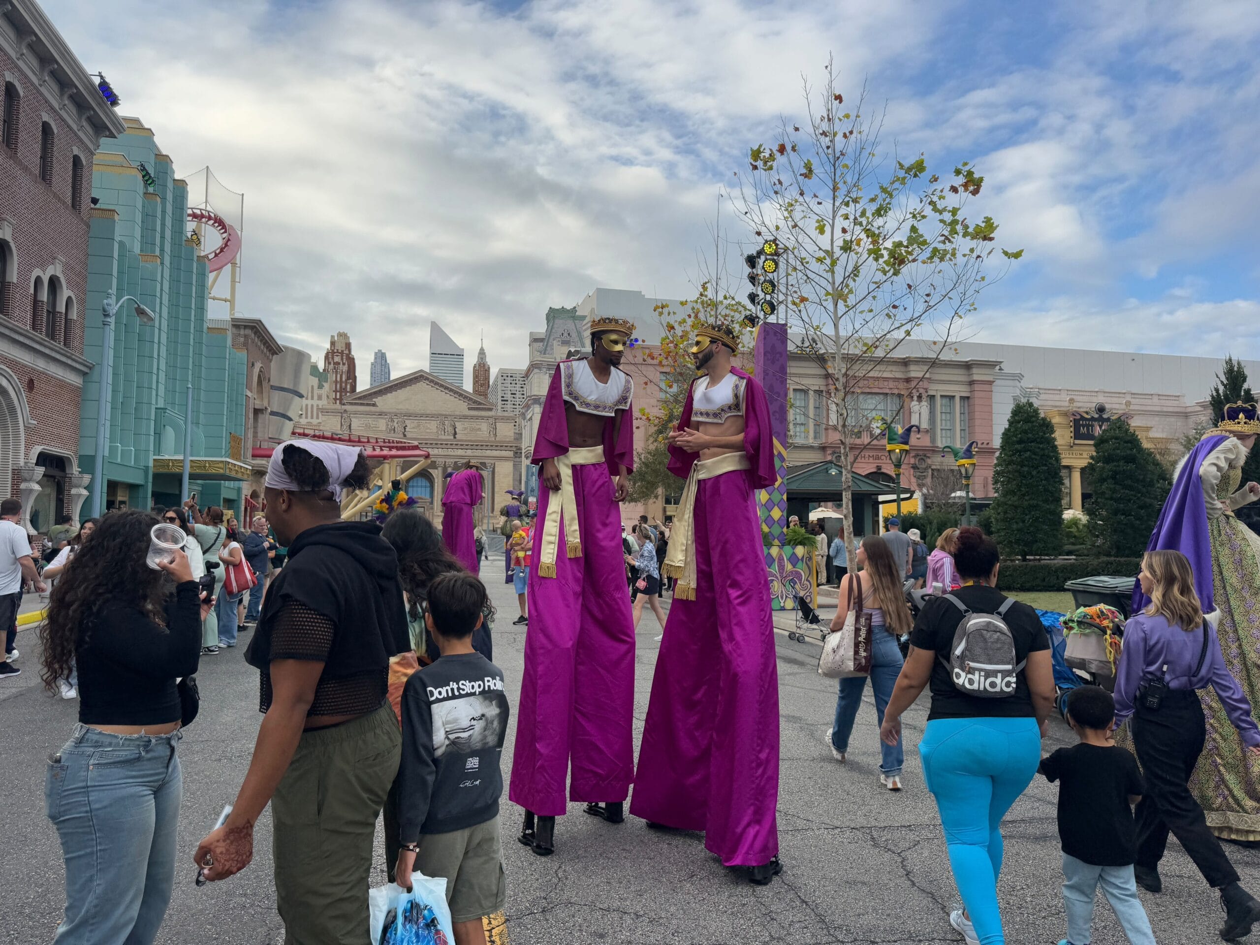Two performers on stilts in vibrant purple costumes captivate the crowd during the Universal Mardi Gras Parade 2025, set against a backdrop of charming buildings and lush trees.