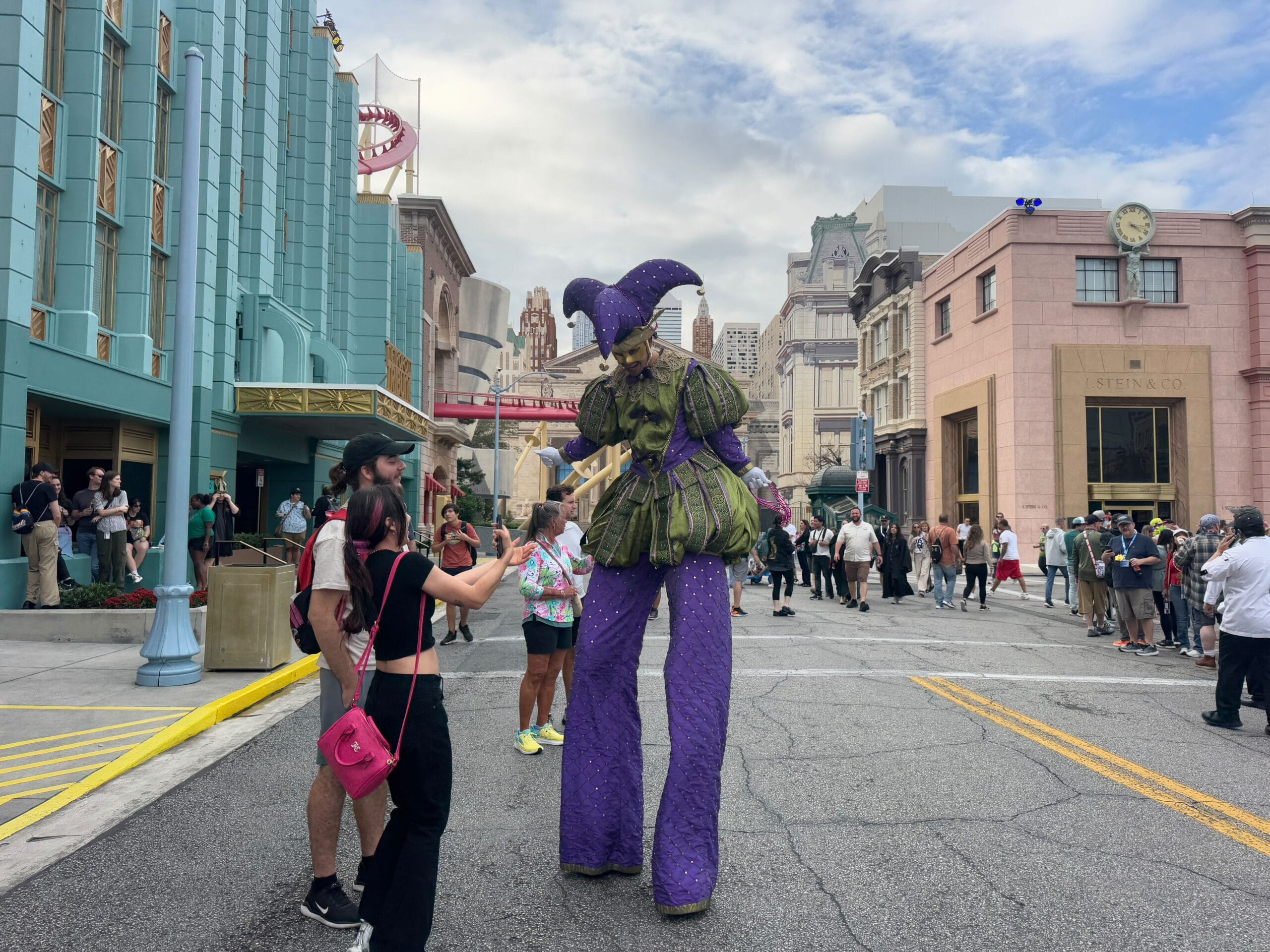 A performer dressed as a jester on stilts captivates the crowd during the Universal Mardi Gras Parade, interacting with guests as they snap photos and record video of this lively theme park street scene.