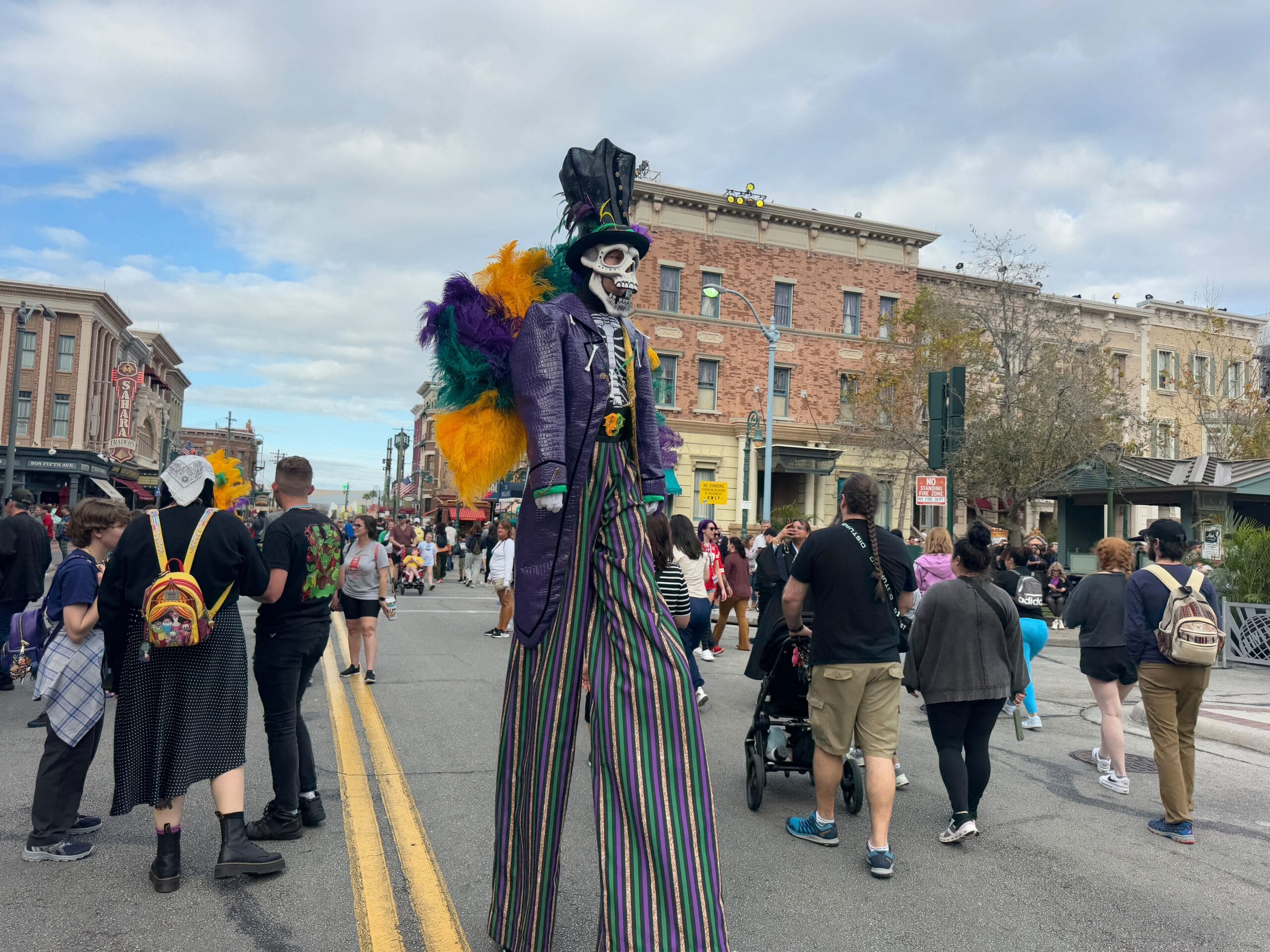 Amid the vibrant Mardi Gras Parade, a person on stilts in a colorful costume and face paint stands tall, captivating the crowd at this universal celebration. The lively energy of the 2025 festival sweeps through the bustling street as people joyfully walk around.