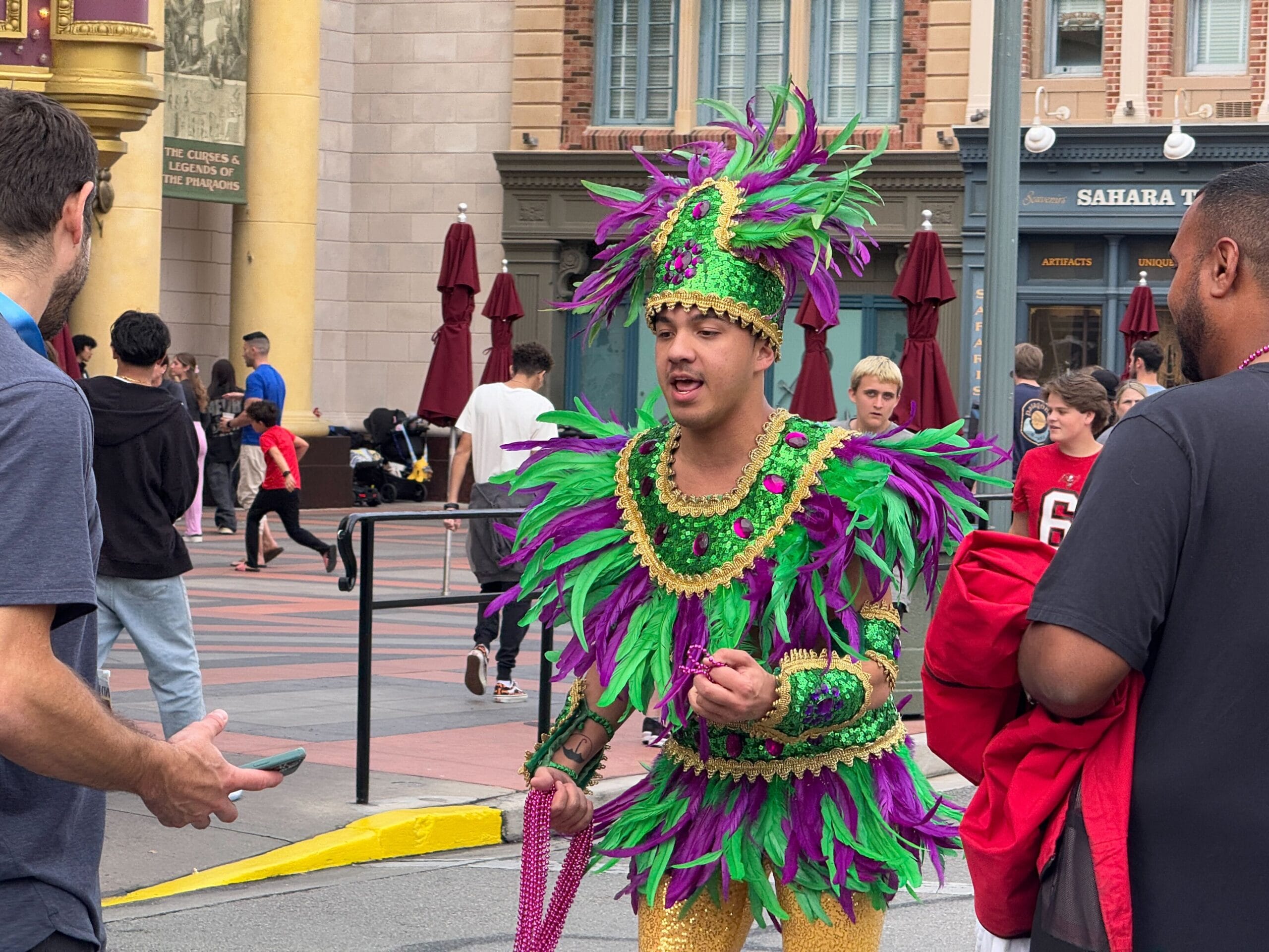 A person in a green and purple feathered costume, holding beads, is captured on video amidst the lively crowd at the 2025 Universal Mardi Gras Parade.