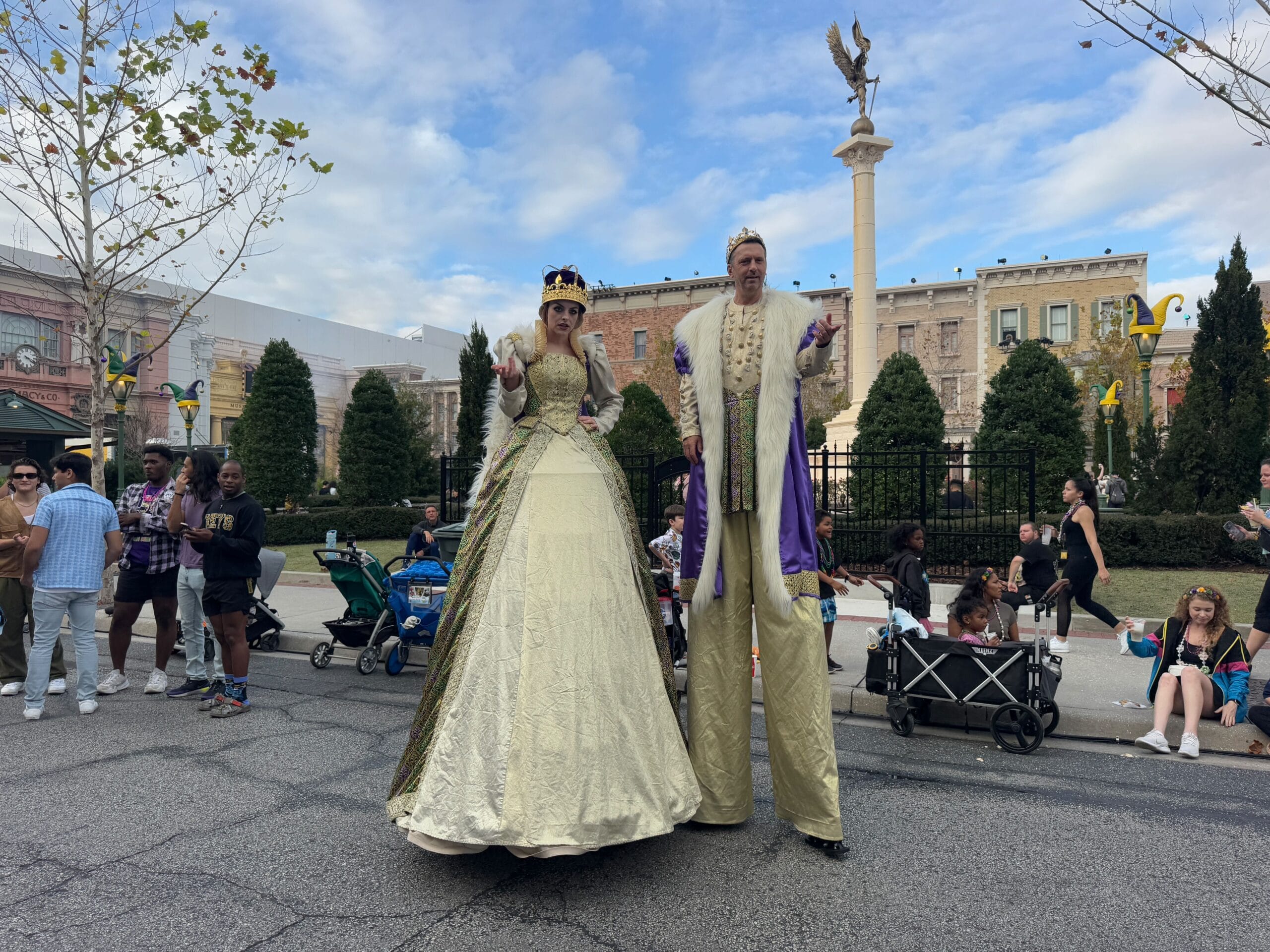 Two people dressed as royalty on stilts strike a pose for photos at the 2025 Universal Mardi Gras Parade. In the lively outdoor setting, a stroller and eager onlookers can be seen in the background.