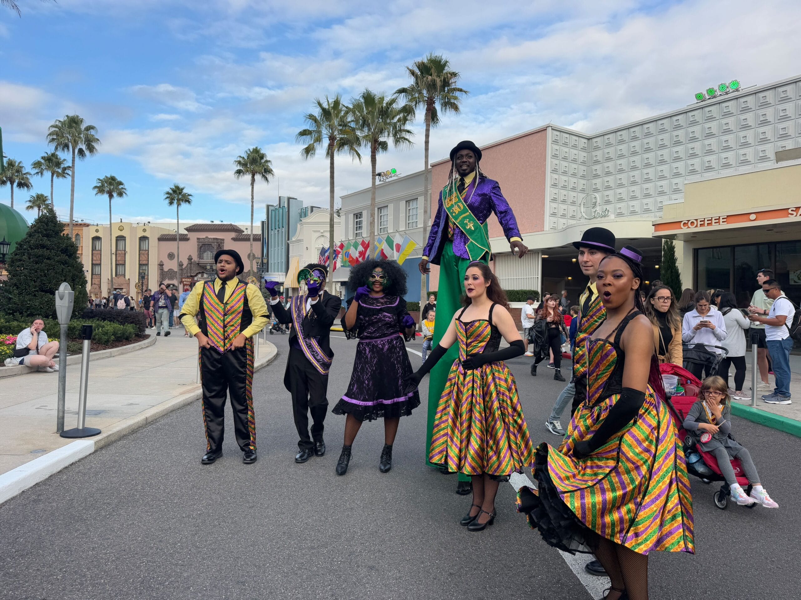 A lively group of performers in vibrant costumes poses on the street during the Universal Mardi Gras Parade 2025. With palm trees and buildings as their backdrop, some don hats and intricate patterns, while spectators capture photos of the colorful spectacle unfolding before them.