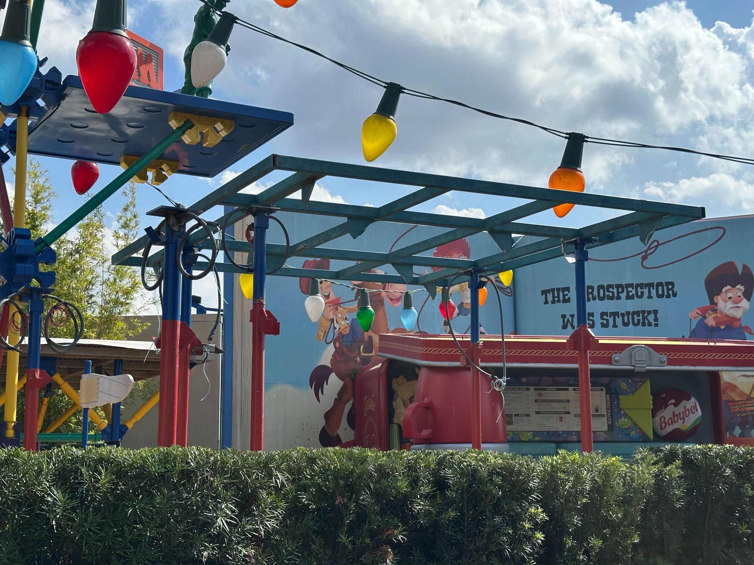 Playground area with large colorful light bulbs, toy structures, and a backdrop featuring cartoon characters and the text "The Prospector Was Stuck." Shrubs are in the foreground.