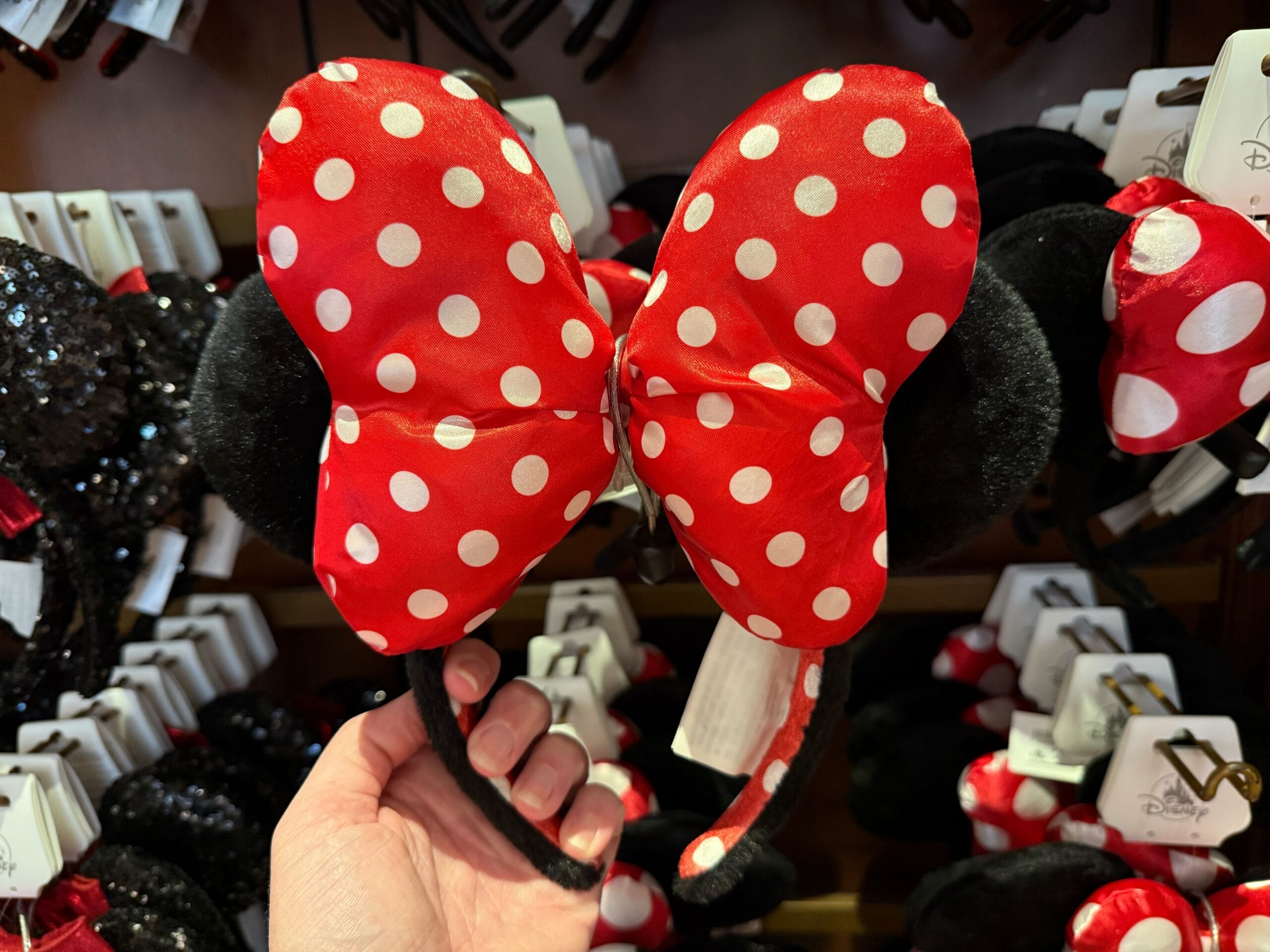 A hand holds a headband with a red polka-dotted bow and black mouse ears, surrounded by similar items displayed on a shelf.