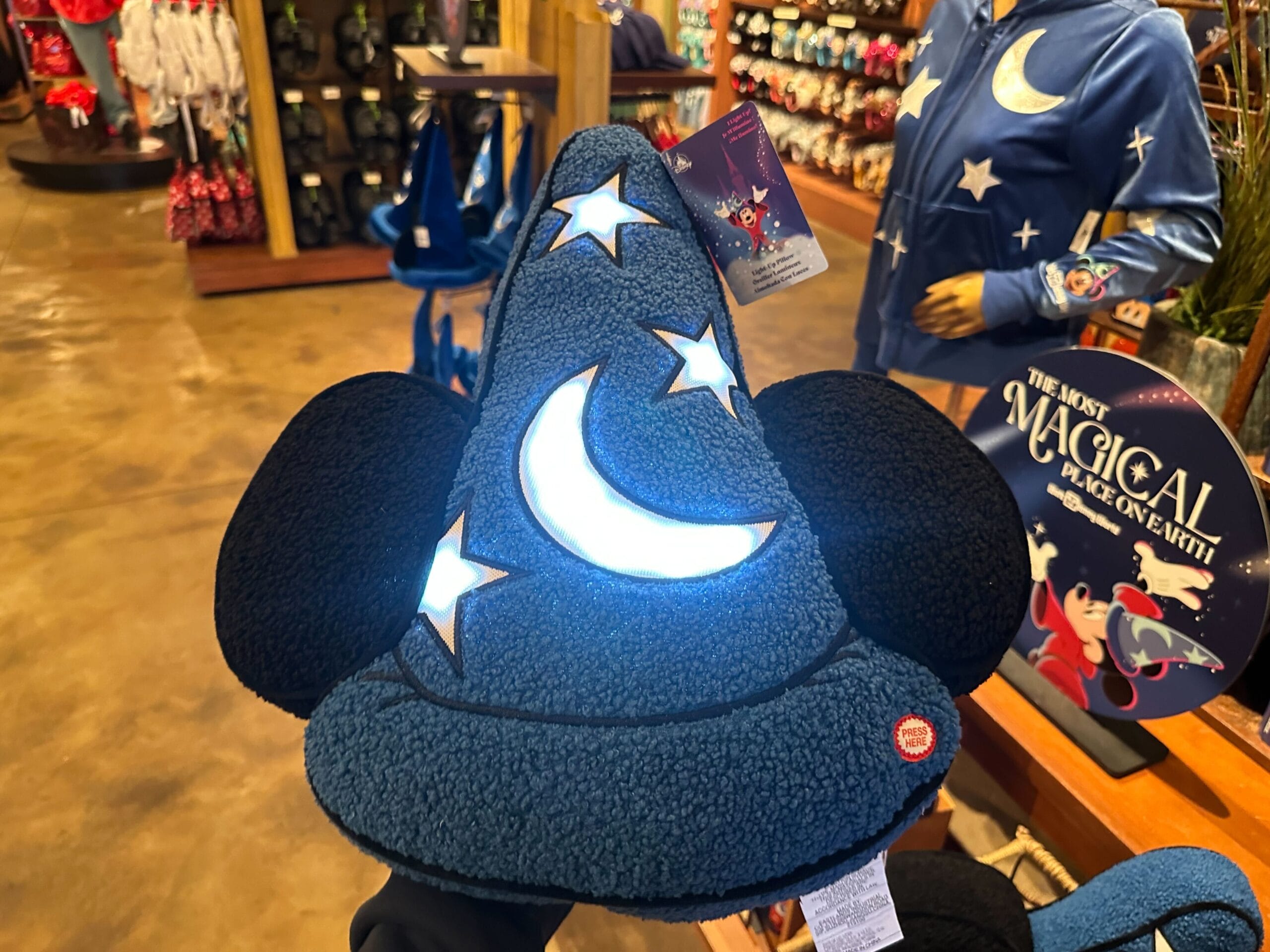 Blue wizard hat with moon and stars pattern, featuring black mouse ears. Displayed in a store with other themed merchandise in the background.