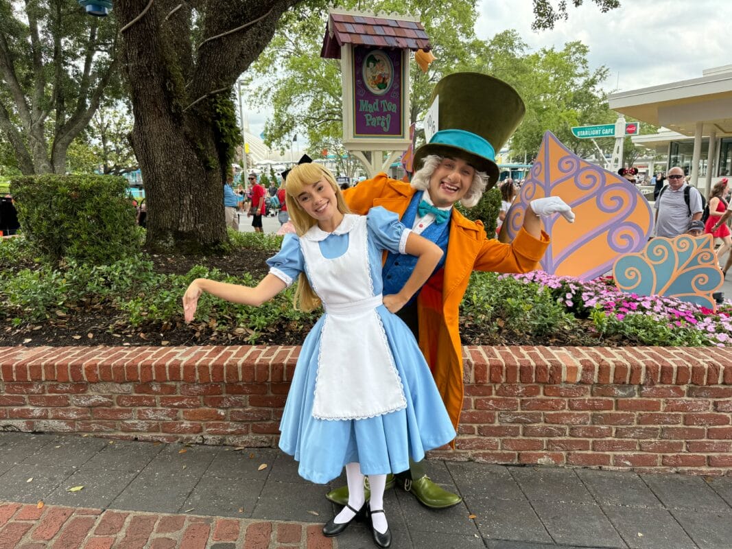 Two people dressed as Alice and the Mad Hatter pose playfully by the Mad Tea Party sign at an amusement park.