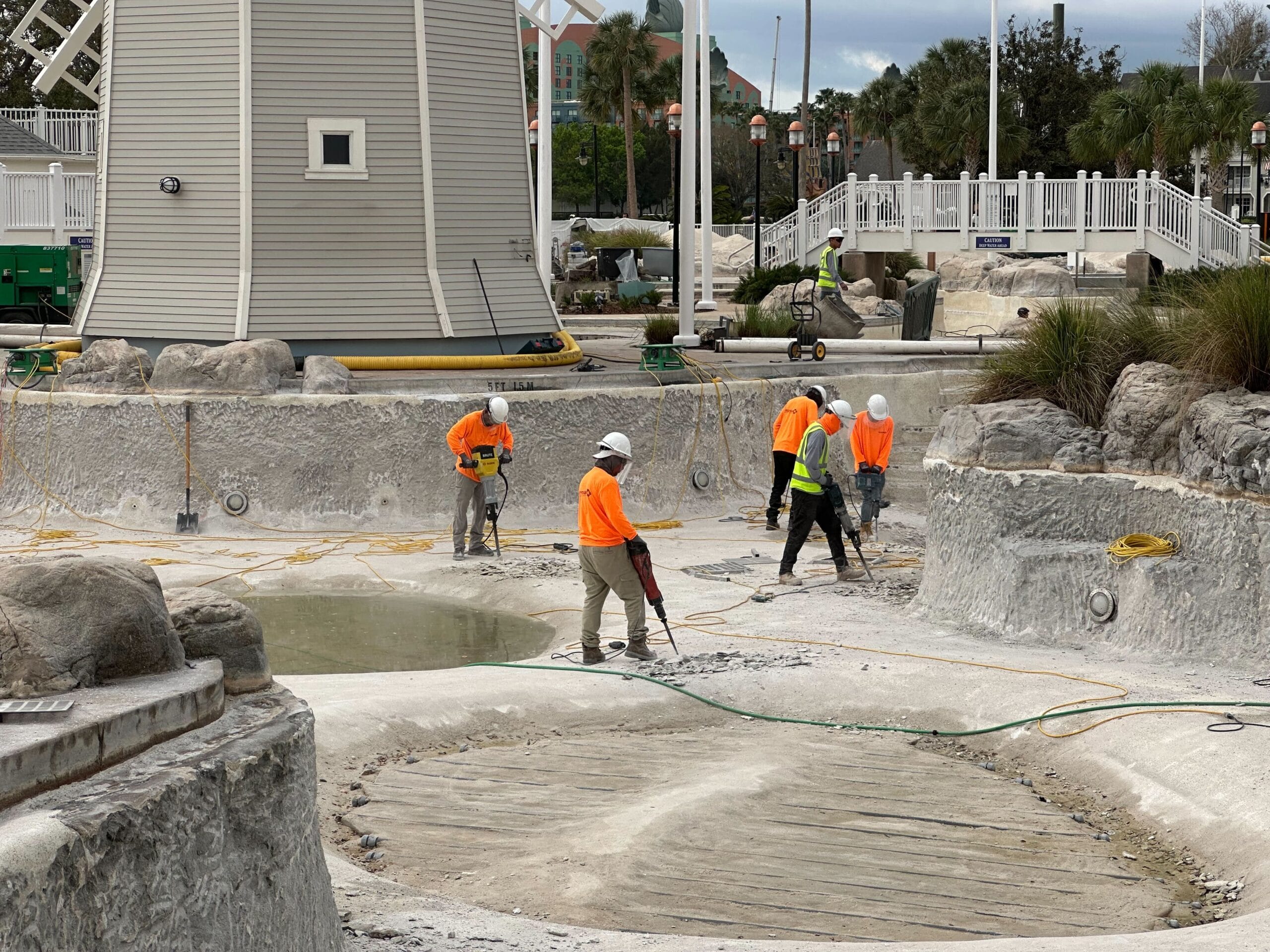 Workers in orange vests and hard hats use equipment to renovate an empty swimming pool area near a small lighthouse structure.