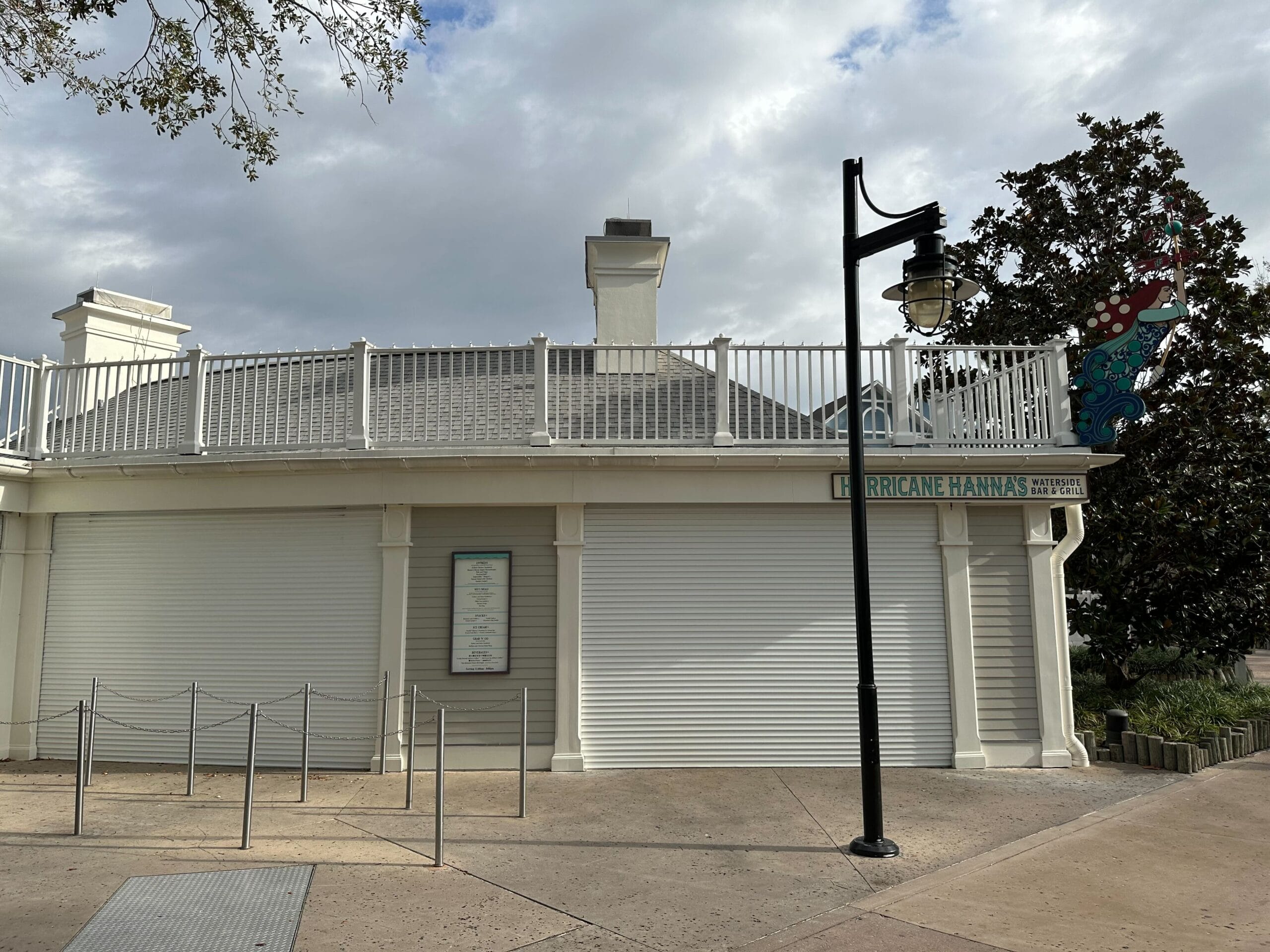 A white building with closed metal shutters, labeled "Hurricane Hanna's," with a lamppost and cloudy sky above.