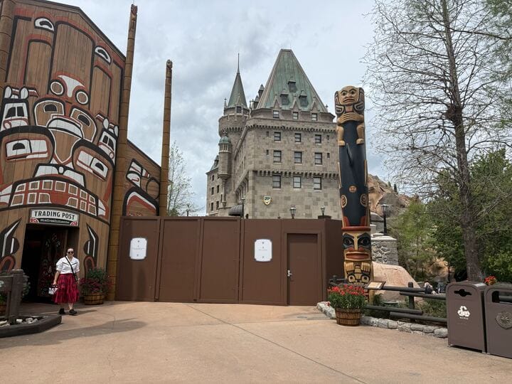 A building adorned with painted designs stands beside a tall wooden totem pole, while a castle-like structure looms in the background. The DVC Welcome Home Center now boasts unobstructed views with scaffolding removed, though construction walls and a trash bin remain in the foreground.