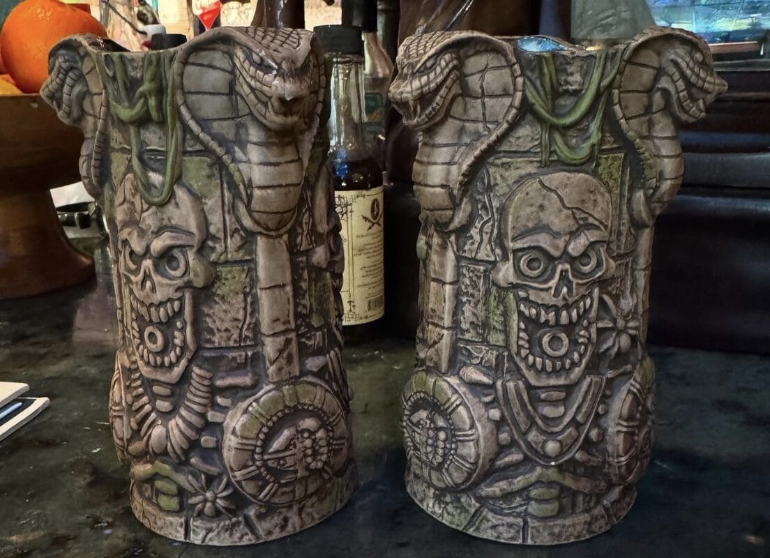 Two intricately carved stone-like statues featuring skulls, skeletal figures, and snakes, with a weathered appearance reminiscent of Indiana Jones Adventure Anniversary props, are displayed on a dark surface.