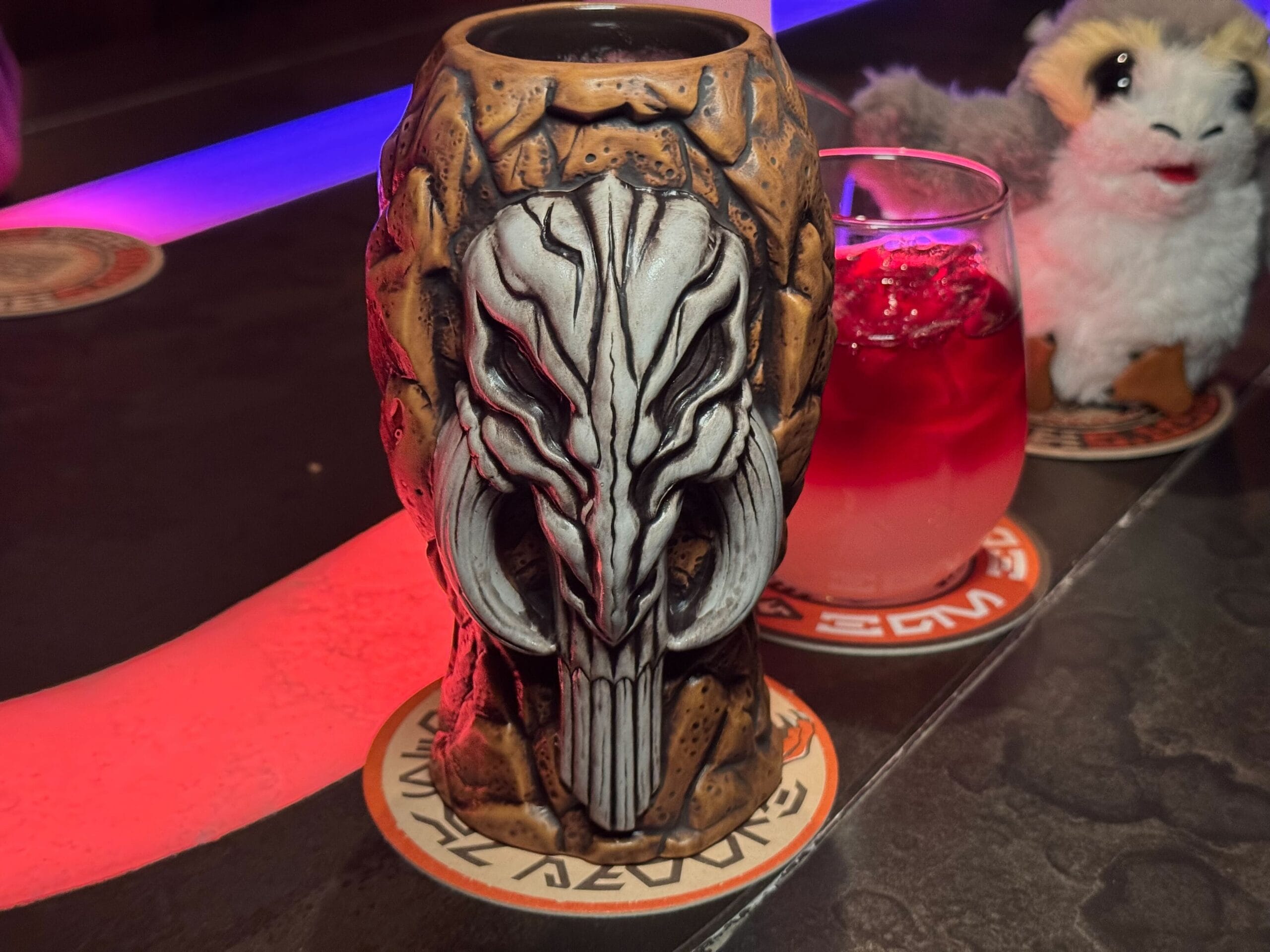 A Mythosaur Mug, resembling a mask, sits on a coaster beside a red drink and a stuffed toy on a dimly lit table adorned with colorful lights.