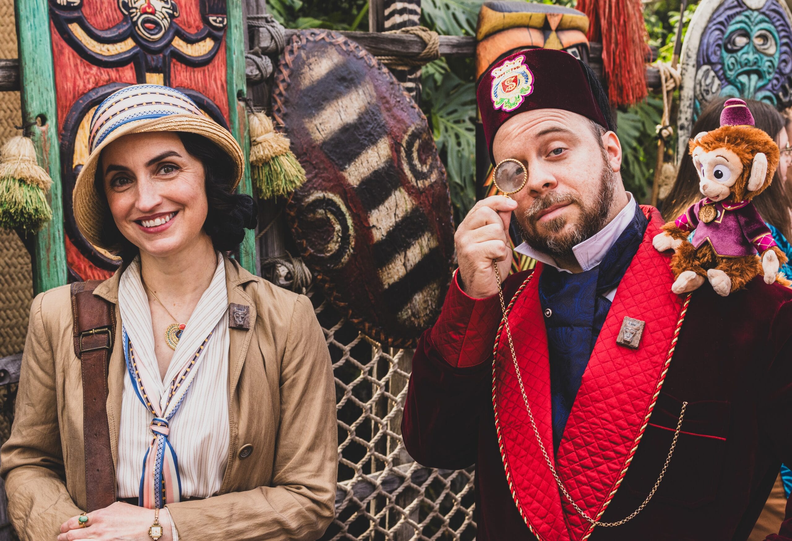Two people dressed in vintage explorer costumes exude creativity as they embark on Adventureland Day 2025. One wears a hat and blazer, while the other sports a fez with a toy monkey and monocle. The decorative, tribal-themed backdrop hints at the magic of Disneyland adventures.