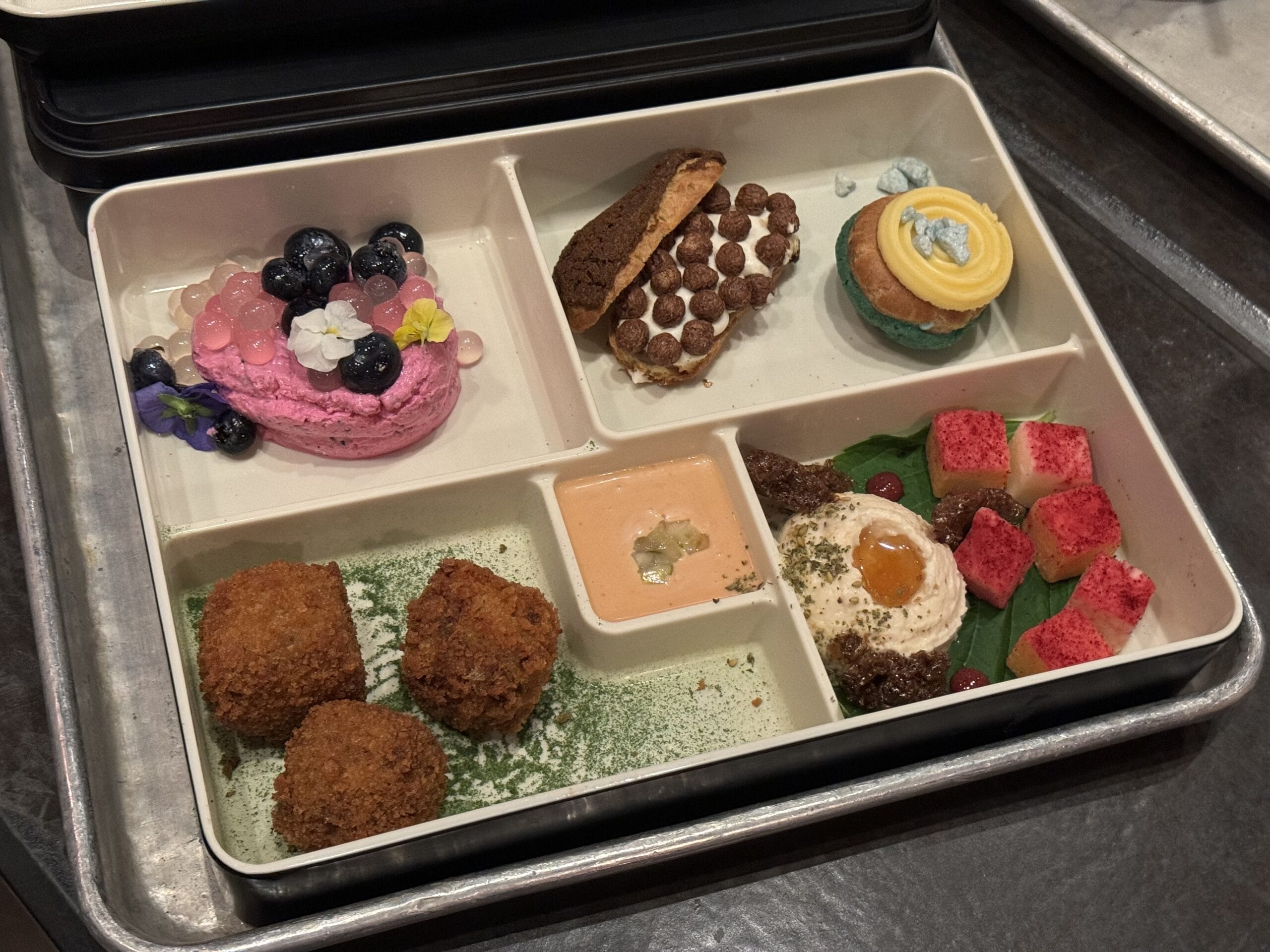 A divided tray featuring delights worthy of a Star Wars dessert party: purple macaron, cookie sandwich, cupcake, savory arancini with sauce, and egg on greens alongside intriguing red cubes.