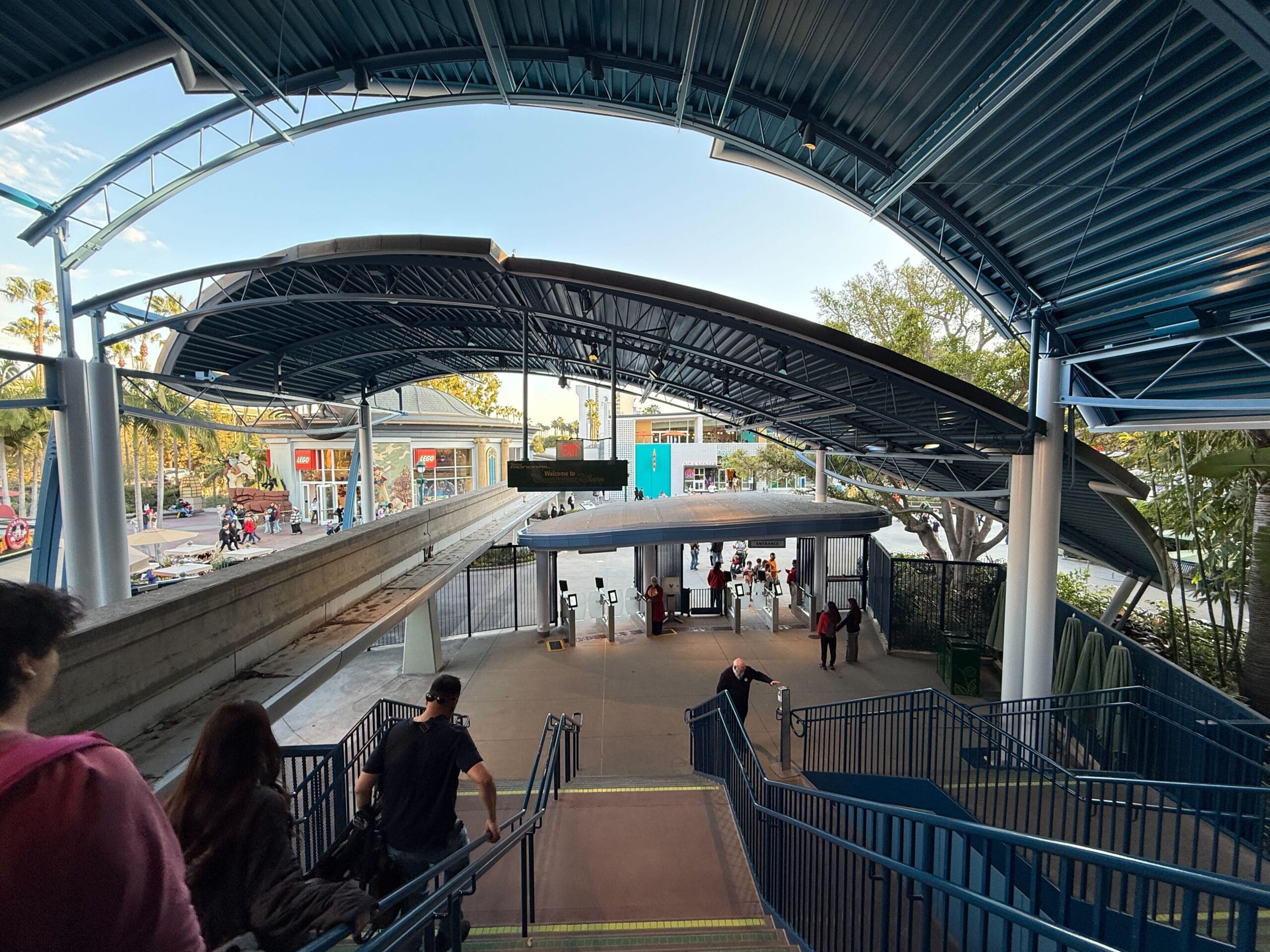 People descending stairs to a bus station platform with a covered waiting area; trees and a street with cars are visible in the background, reminiscent of the orderly flow at the entrance to the Disneyland Monorail.