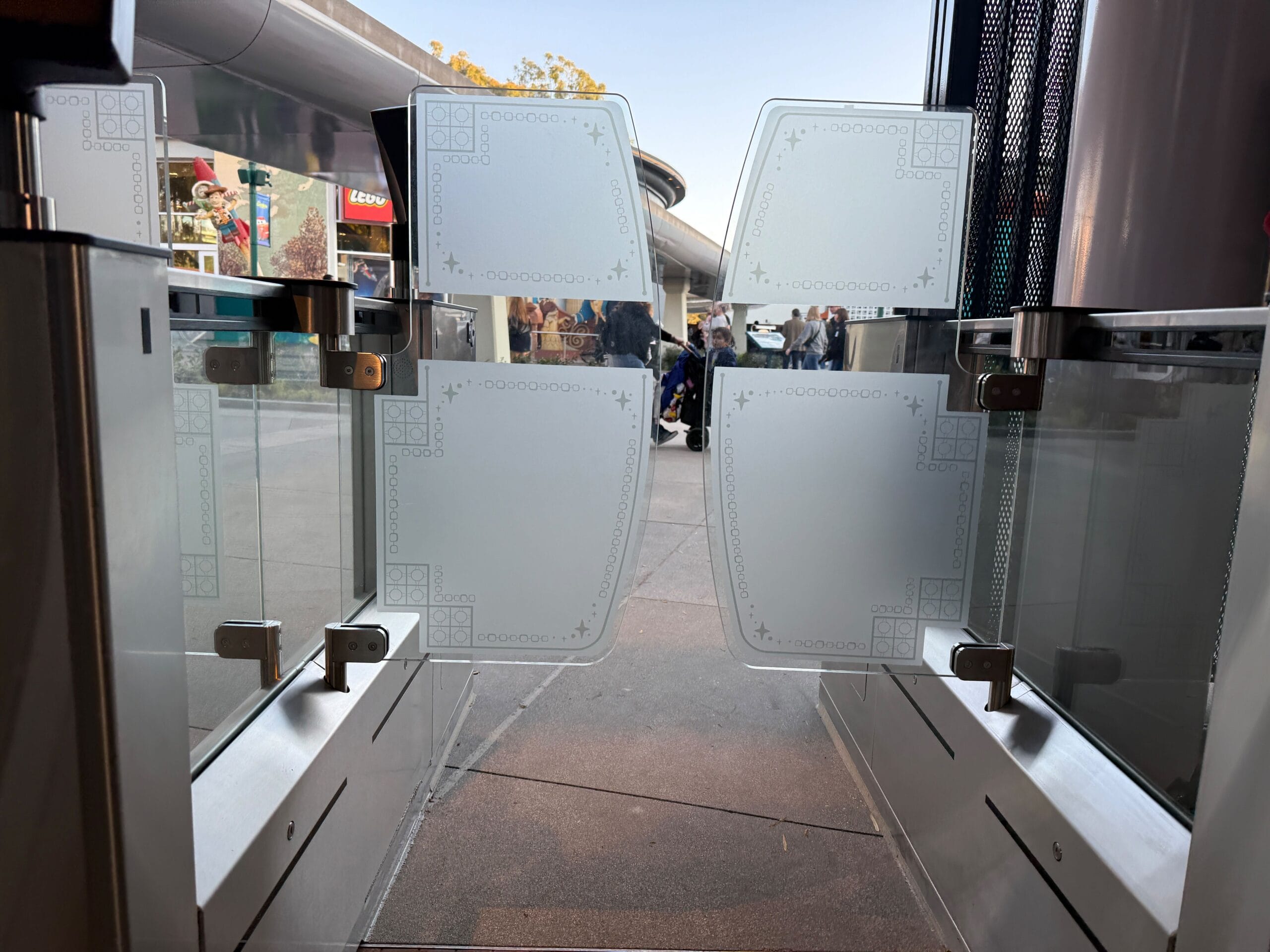 The frosted glass gates with decorative patterns slide open, unveiling a scene bustling with people and shops, reminiscent of the enchanting journeys beyond the gates of the Disneyland Monorail.
