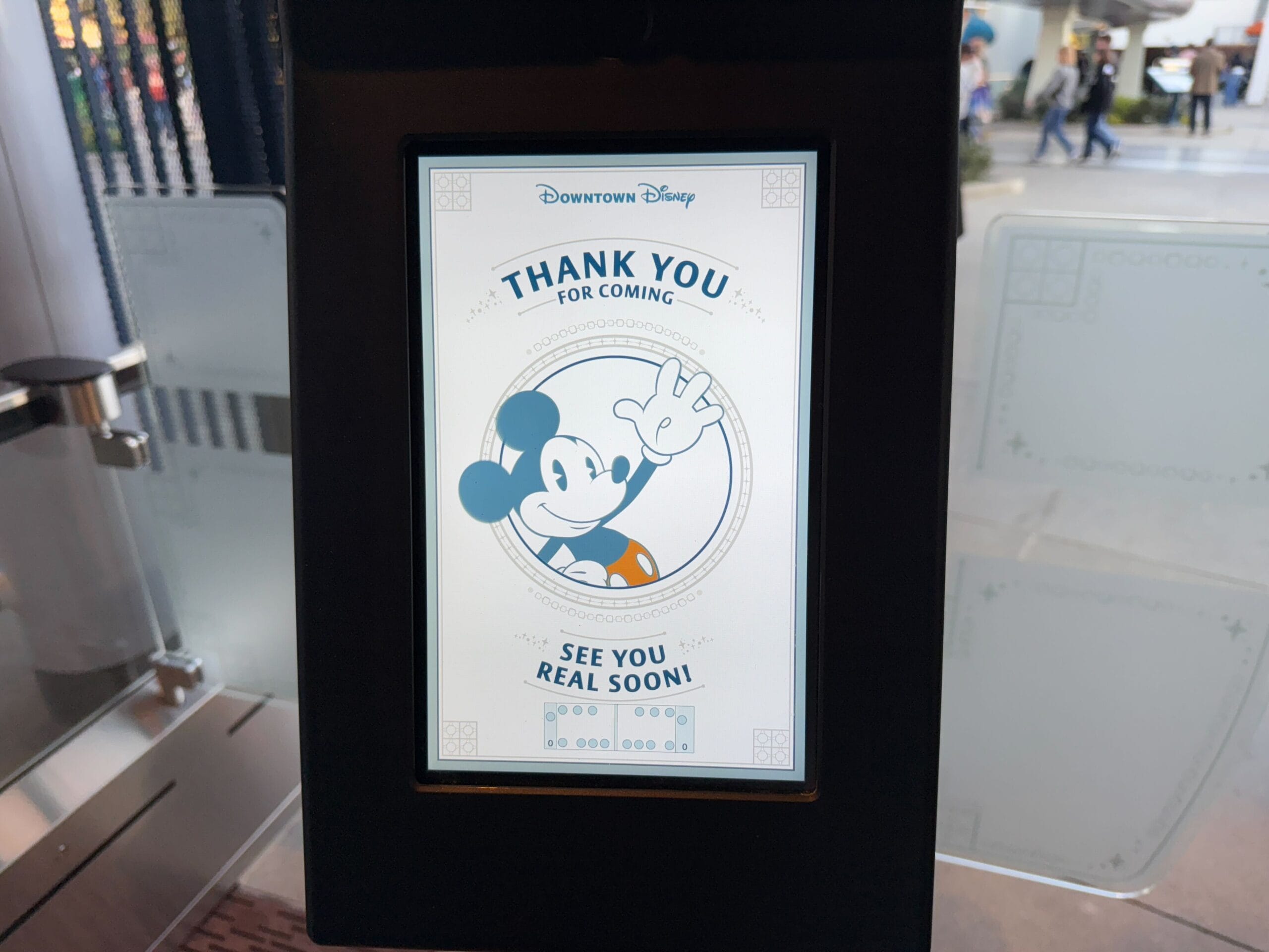 A screen at Downtown Disney displays a message with a cartoon character waving, saying "Thank you for coming" and "See you real soon!" as the Disneyland Monorail glides smoothly in the background.