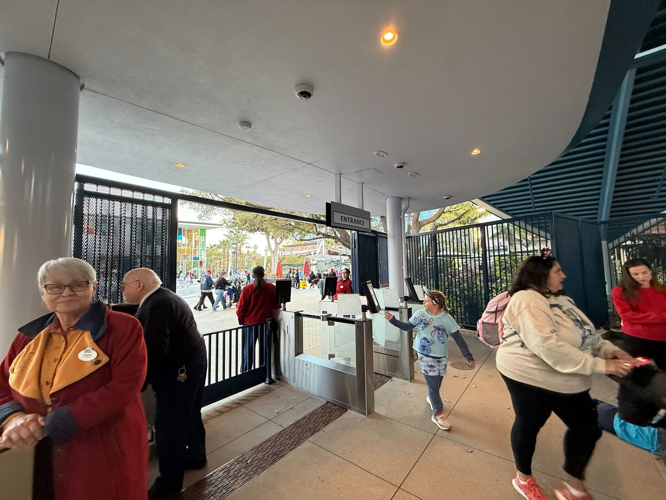 People walking through a security checkpoint with metal detectors at the entrance to the Disneyland Monorail. A woman in a uniform stands to the side, ensuring everything runs smoothly for visitors eager to embark on their magical journey.