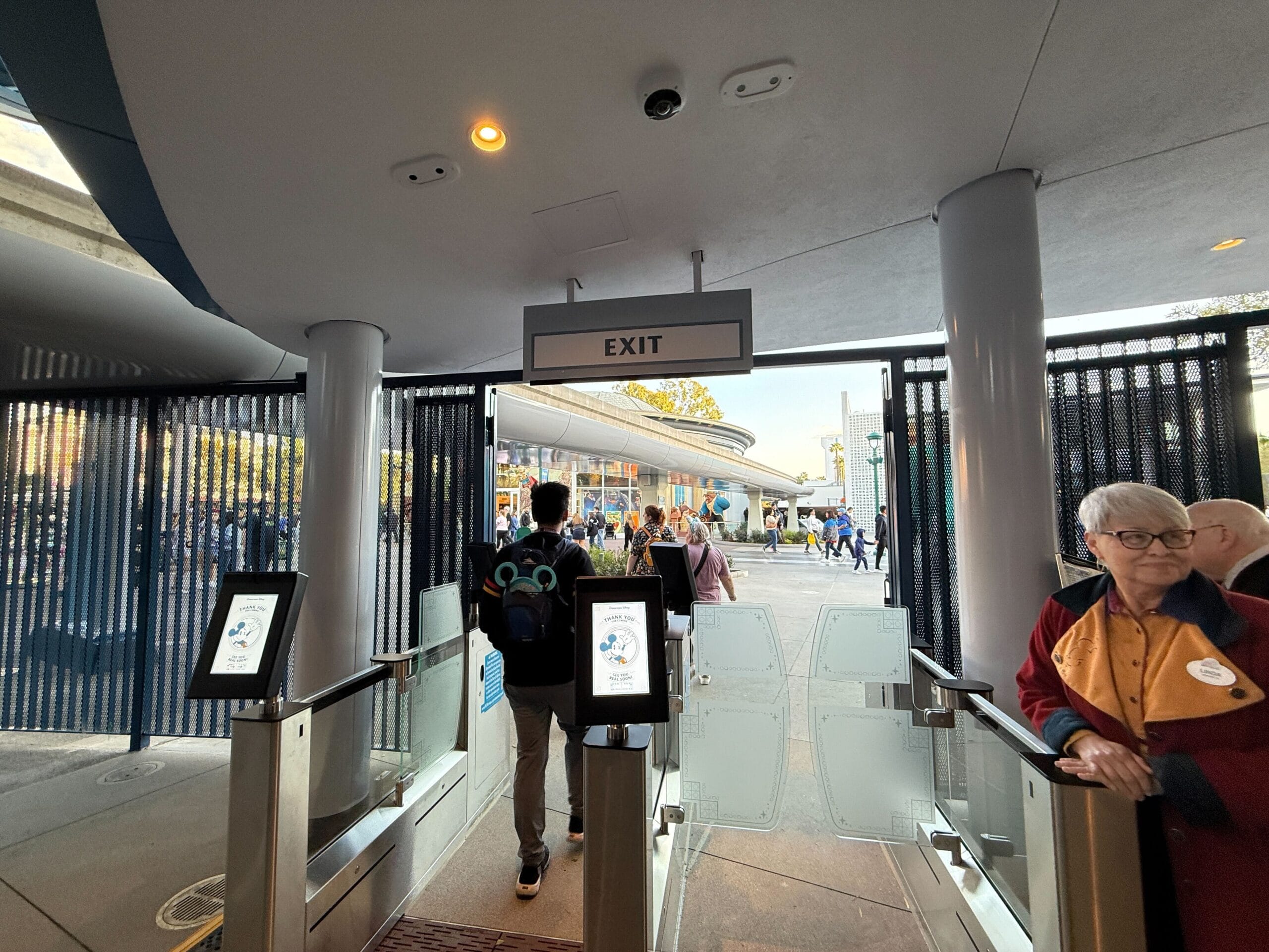 People exiting a building through automatic gates, with an exit sign overhead, look like they could be boarding the Disneyland Monorail. On the right, there's a person in a red and black jacket with a name tag.