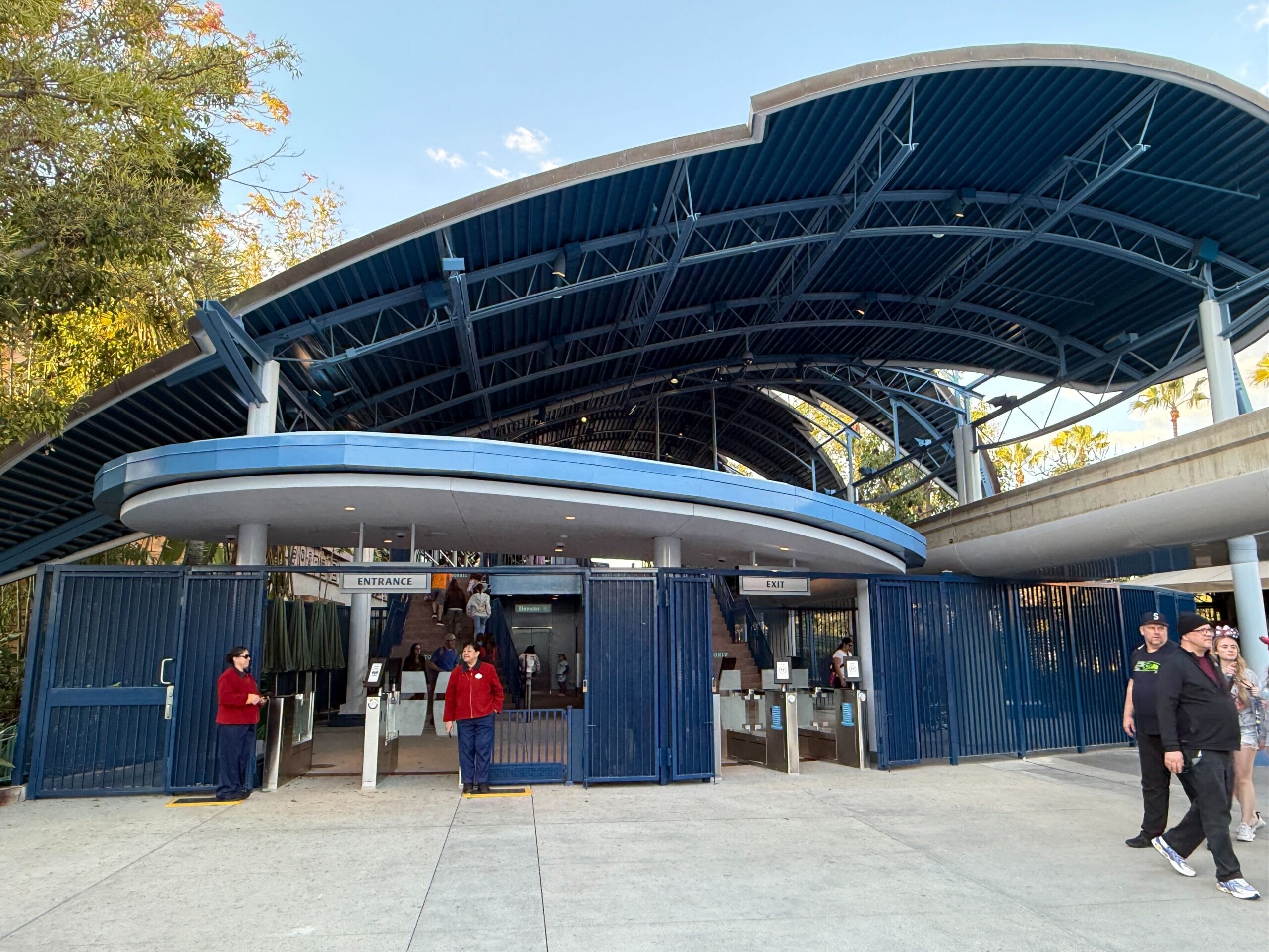 The outdoor entrance gate, featuring a sleek blue canopy and metal turnstiles, welcomes visitors as they wait near the iconic Disneyland Monorail. Lush trees provide a picturesque backdrop.