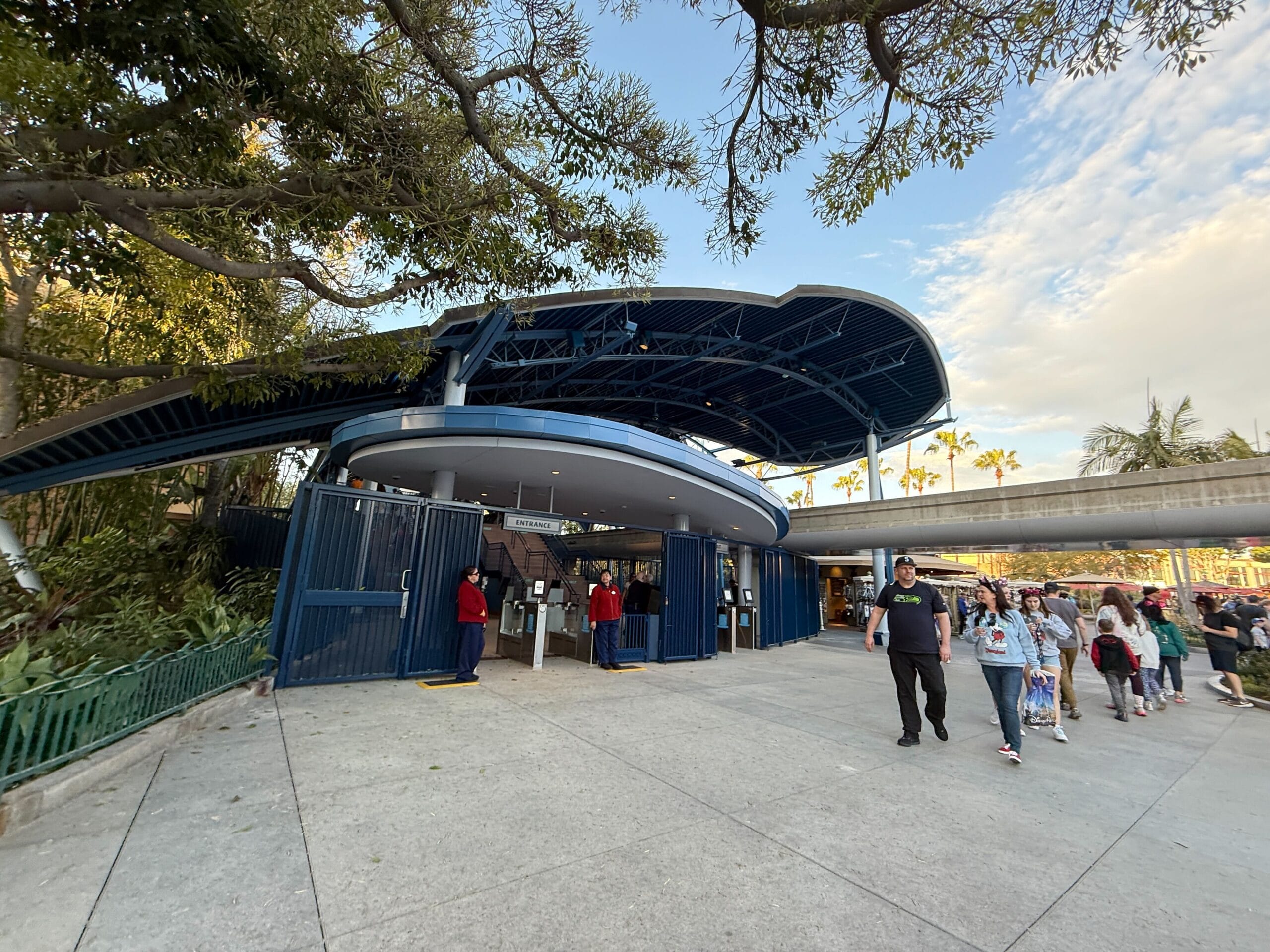People stroll near a large blue and white curved structure, reminiscent of the Disneyland Monorail with its sleek design and entry gates, all framed by trees and a clear sky.