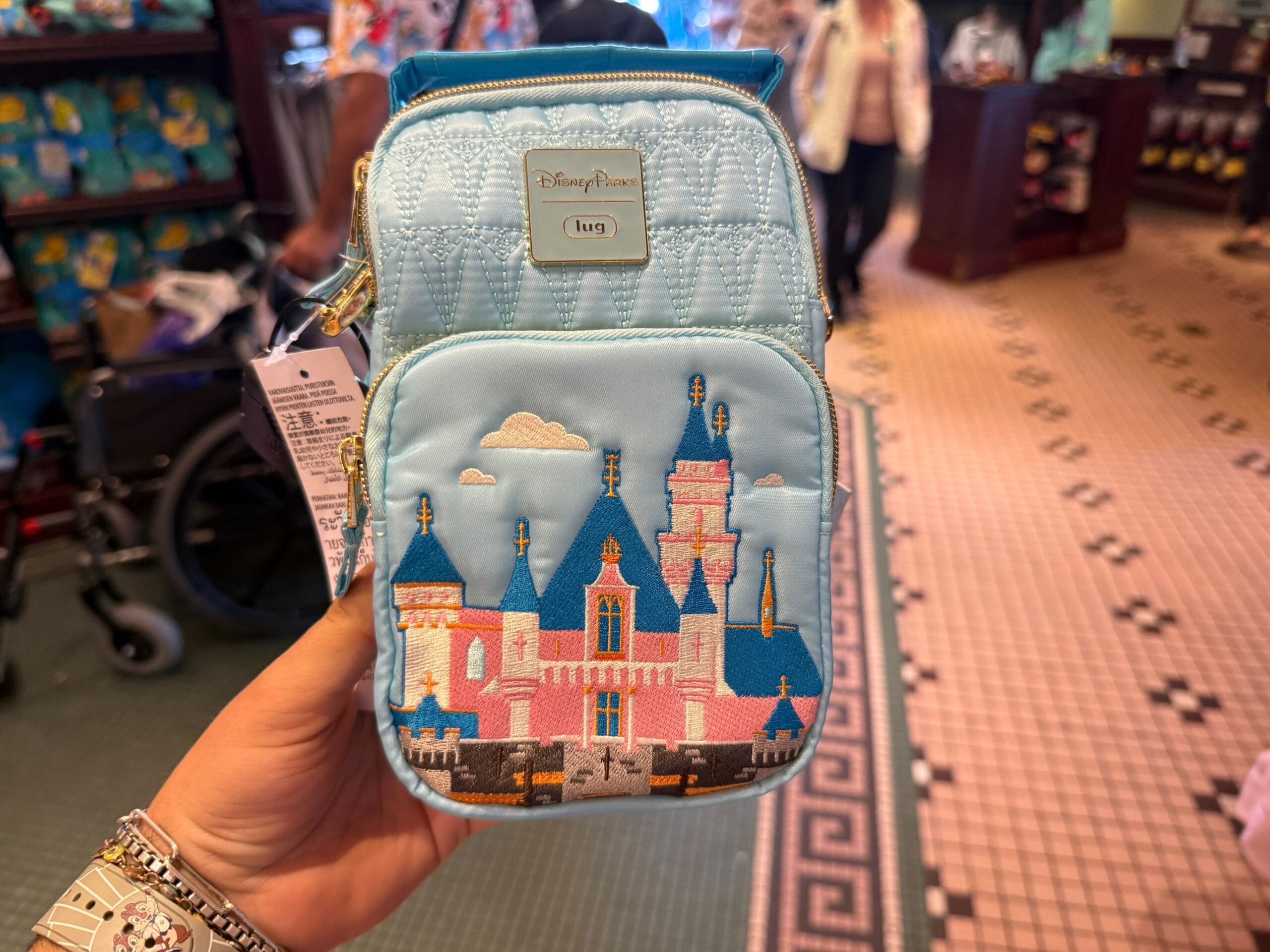 A hand holds a blue Disney-themed bag with a castle design in a store. People and shelves with merchandise are visible in the background.